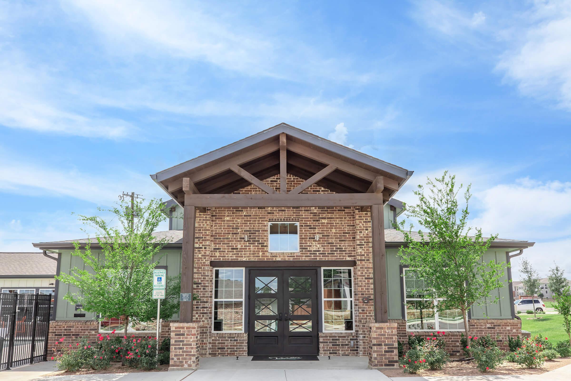 Brick building with large front entrance, featuring a wooden porch and gabled roof. Flanked by green trees and shrubs, under a blue sky with soft clouds. There is a paved walkway leading to the door, and a fence visible in the background.