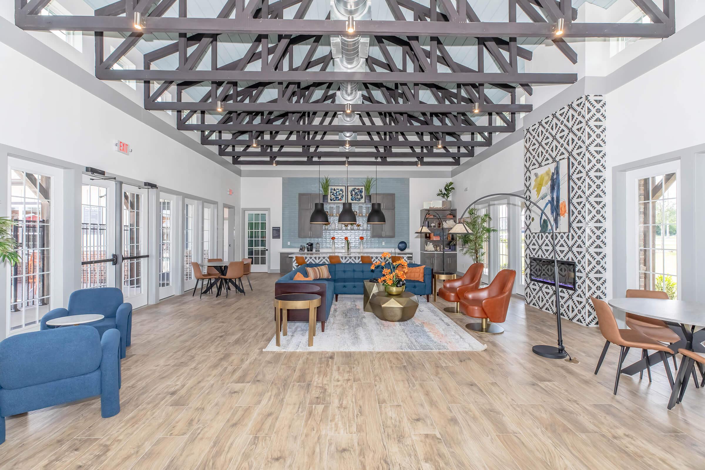 Interior of a stylish lounge area featuring high ceilings with exposed beams. There are comfortable seating options including blue and brown chairs, a large sofa, and a coffee table. A decorative wall with artwork is visible, along with dining tables and a modern kitchen setup in the background. Natural light floods the room through large windows.