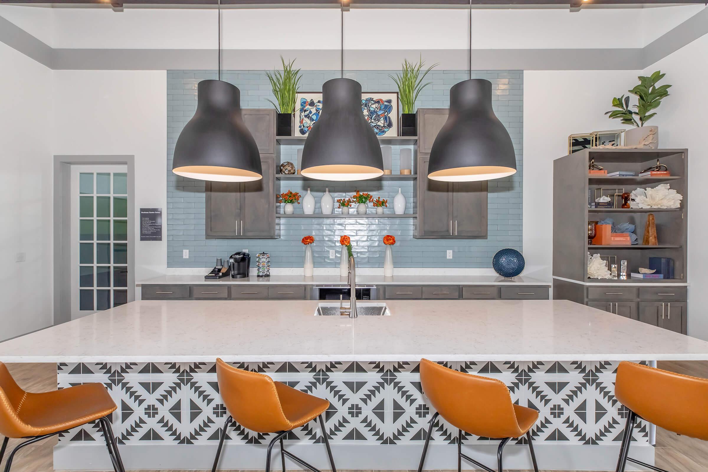 Modern kitchen featuring a large island with a patterned base, three sleek black pendant lights hanging above, and stylish orange bar stools. The backsplash is a light blue tile, complemented by wooden cabinets adorned with decorative plants and colorful decor. Natural light enhances the inviting atmosphere.