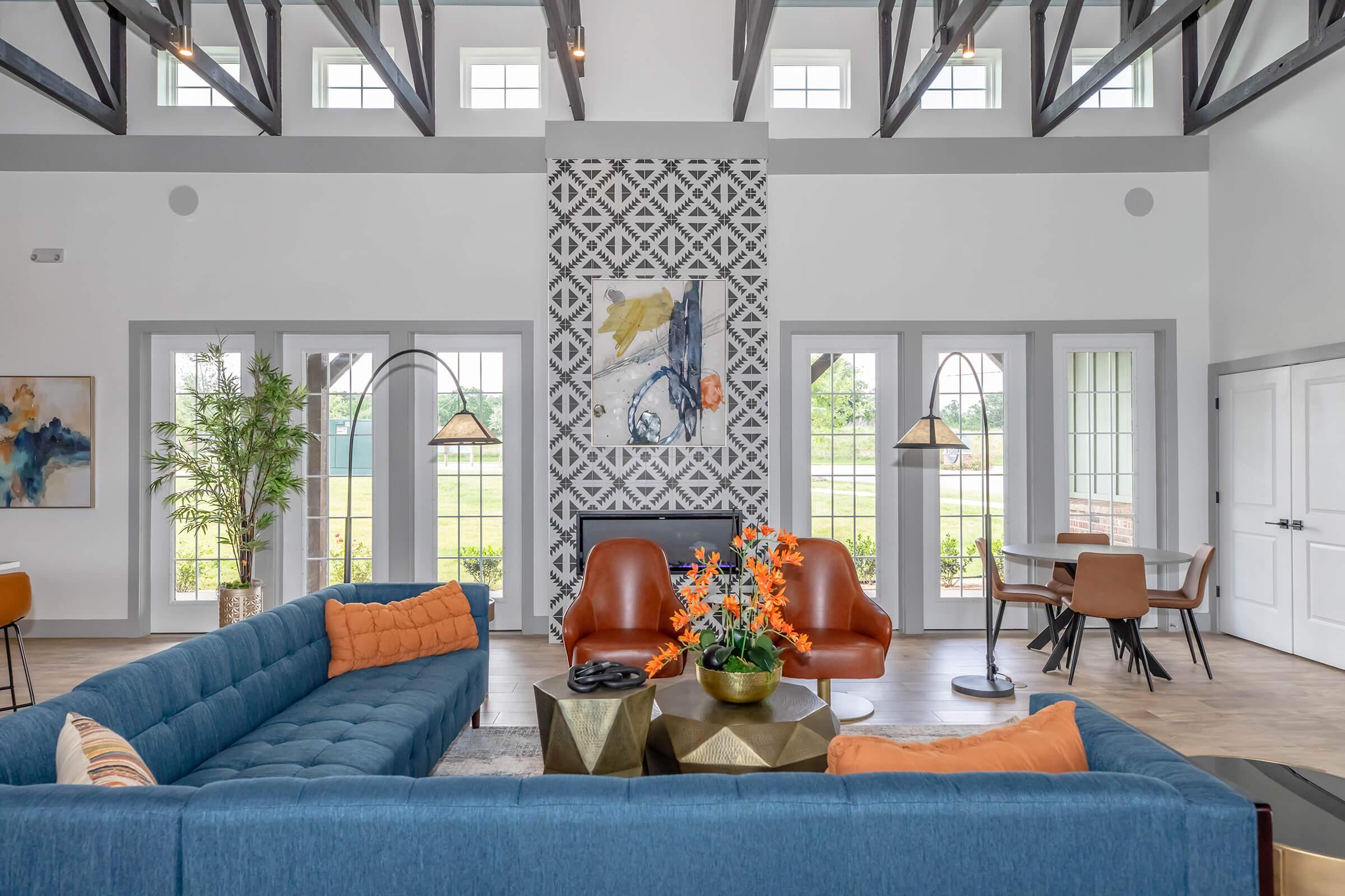 A modern interior of a spacious living area featuring a blue sectional sofa with orange and striped cushions, two brown leather chairs, a coffee table with a decorative plant, and large windows that allow natural light. Decorative artwork and plants enhance the stylish decor.