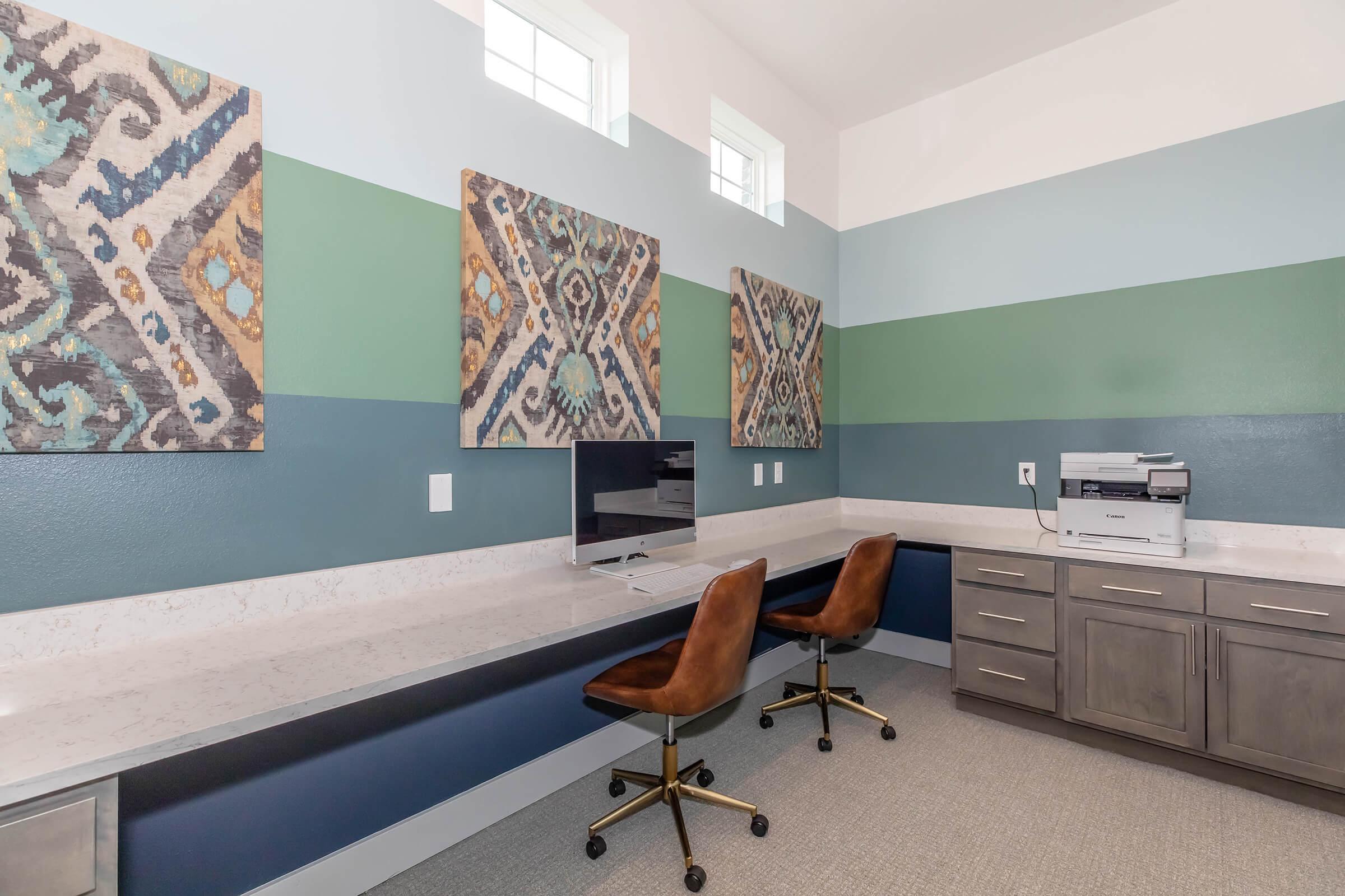 A modern workspace featuring a long countertop with a computer monitor and two rolling chairs. The walls are painted in alternating shades of green and blue, adorned with two decorative wall hangings. A printer is positioned on the right side of the countertop. Natural light comes through the small windows above.