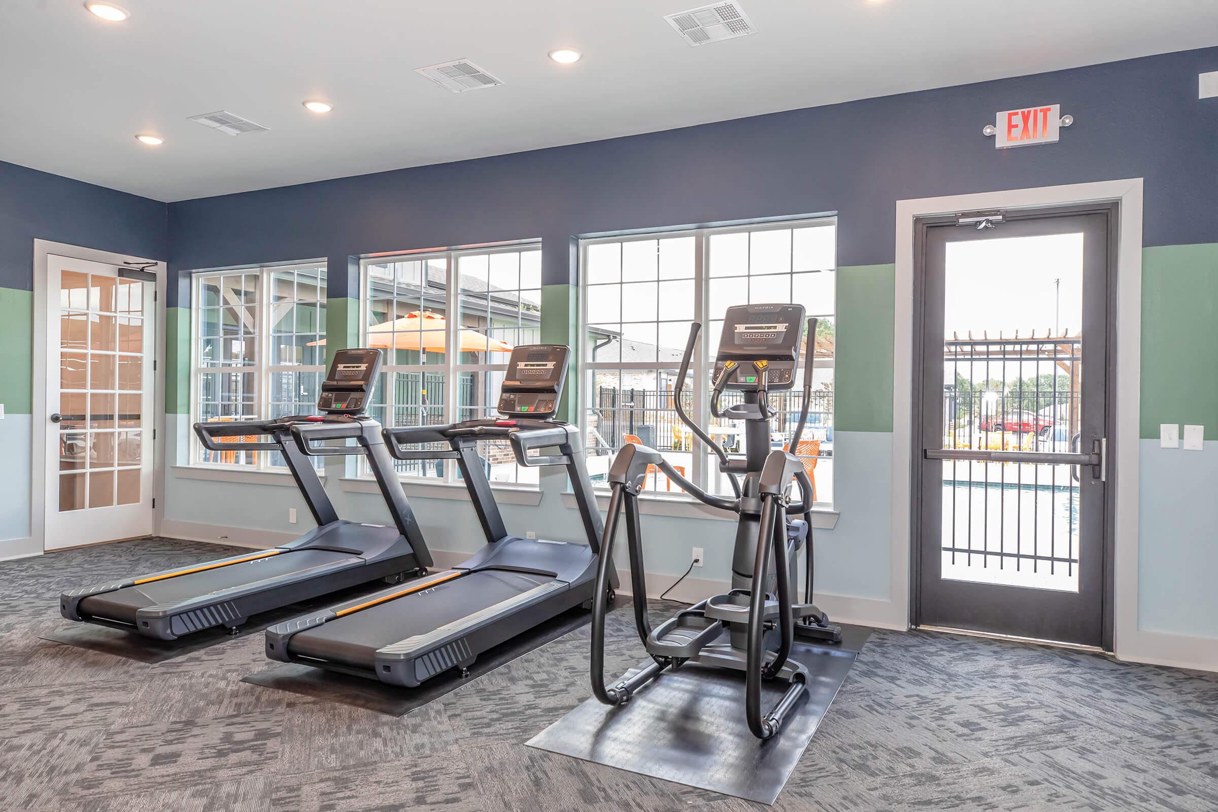 A modern gym with two treadmills and an elliptical machine situated near large windows. The space is well-lit, featuring a door leading outside and access to a pool area. The walls are painted in shades of blue and green, creating a fresh and inviting atmosphere.