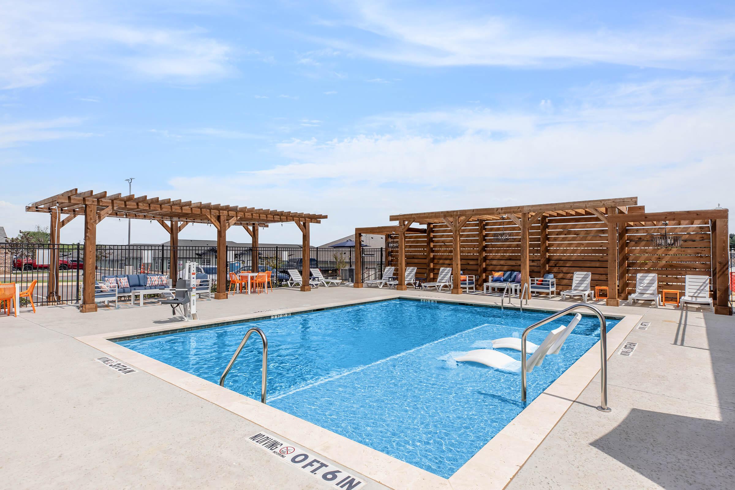 A swimming pool surrounded by lounge chairs and wooden pergolas. The pool features steps leading in and is set against a clear blue sky. There are several empty chairs and a fenced area in the background, suggesting a recreational space ideal for relaxation.