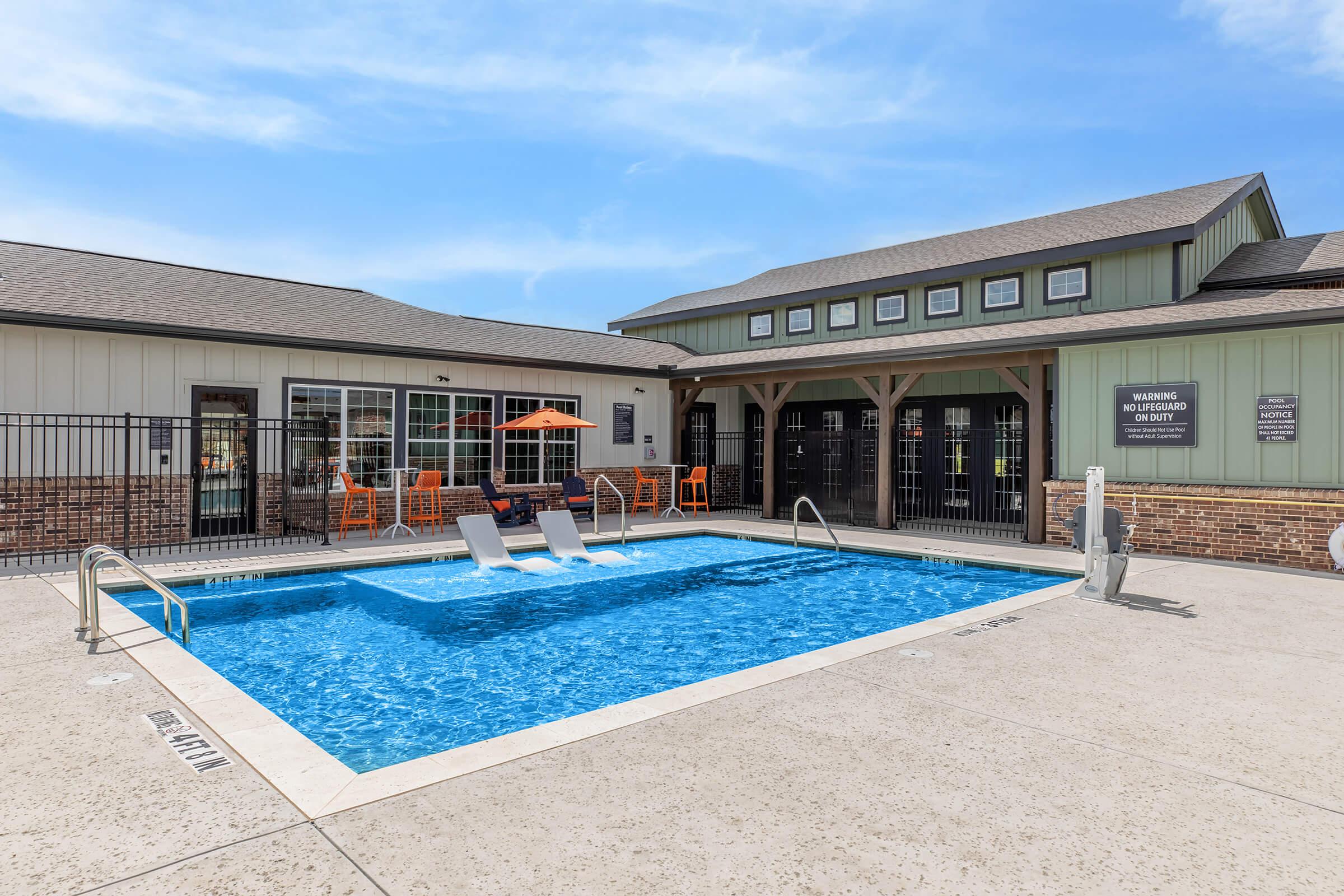 A clear blue pool with two lounge chairs on the deck, surrounded by a fence. In the background, a building features large windows and an awning with orange chairs and tables. The scene is well-lit under a sunny sky, creating a relaxing, inviting atmosphere.