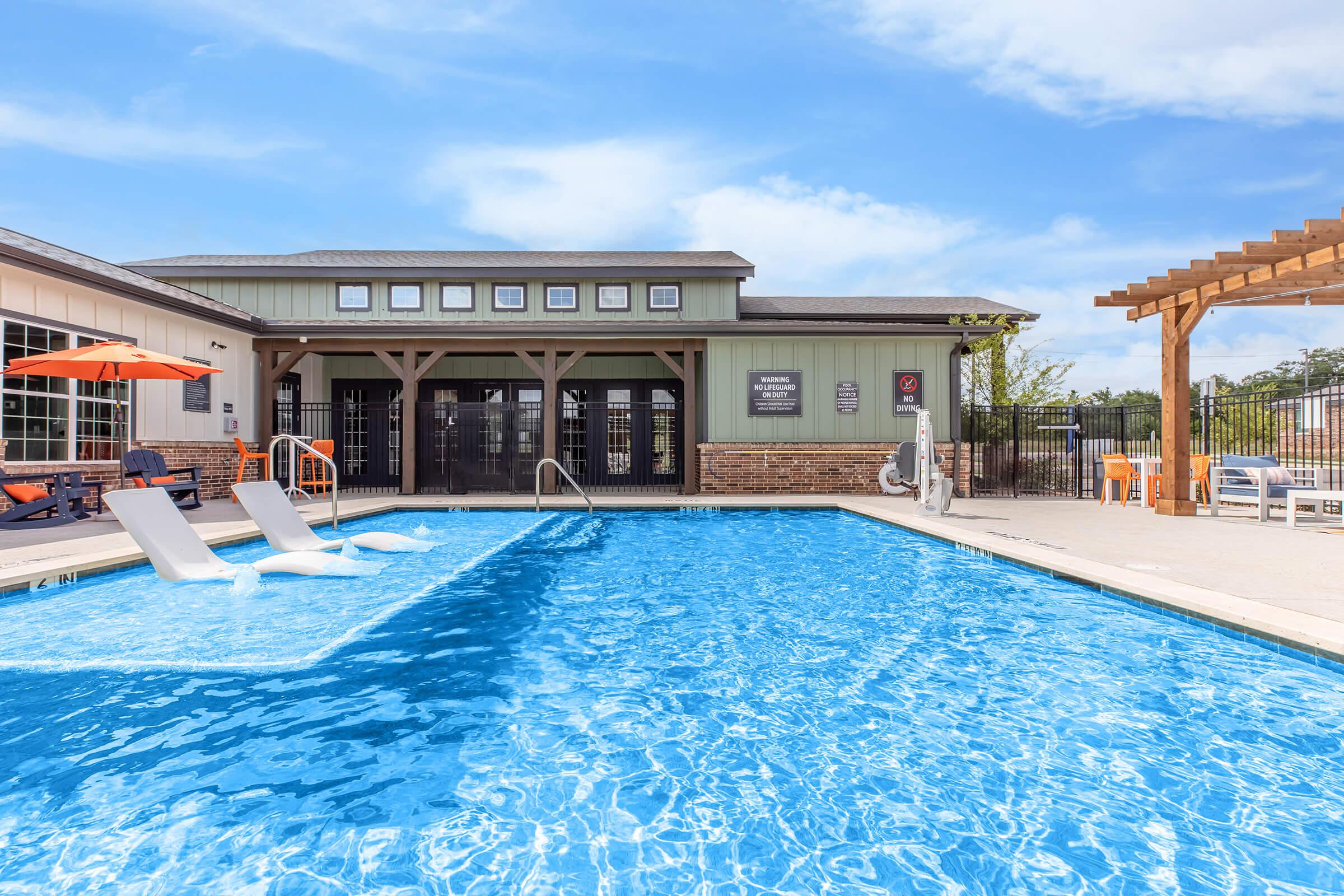 A clear swimming pool with lounge chairs on the side, surrounded by a well-maintained area. In the background, a modern building with large windows and a pergola is visible. The sky is bright blue, adding to the inviting atmosphere of the pool area.
