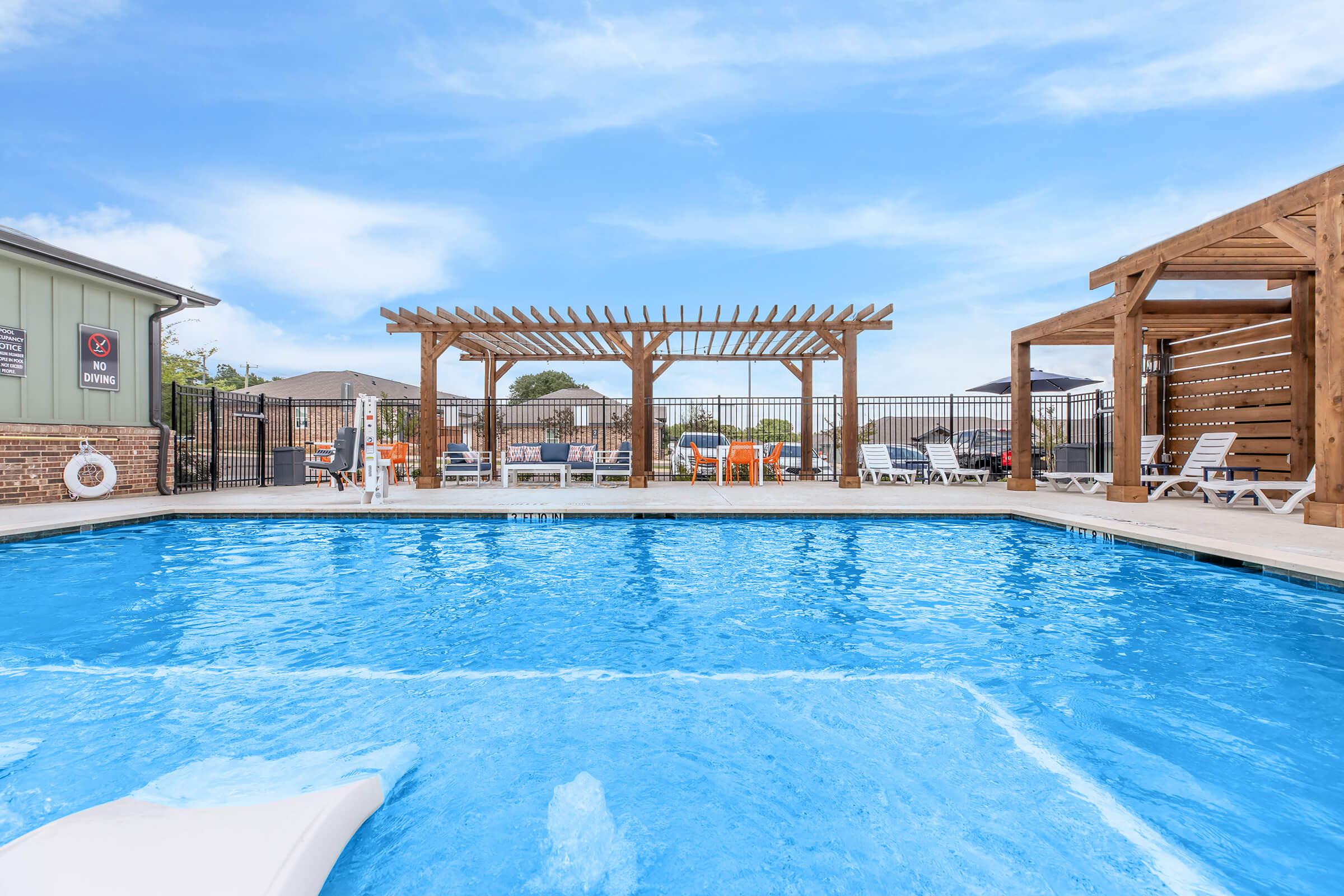 A sparkling blue swimming pool with a wooden pergola in the background. White lounge chairs are arranged around the pool area, and a wooden structure is visible to the right. The sky is clear with a few clouds, creating a bright and inviting atmosphere.