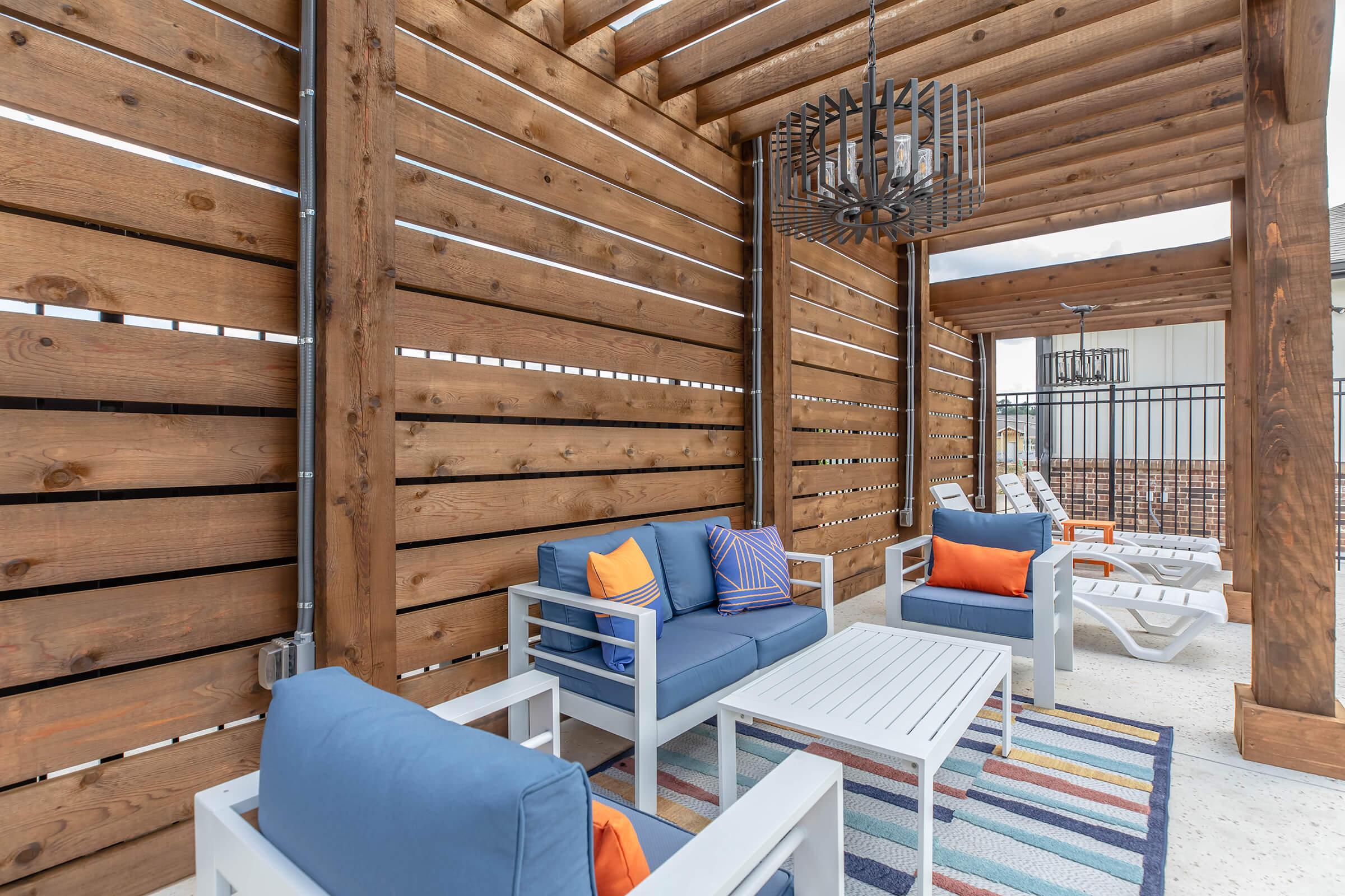A cozy outdoor seating area featuring blue cushioned furniture with orange accents, a white coffee table, and a colorful striped rug. The space is framed by wooden slats and has a decorative chandelier overhead, creating a warm and inviting ambiance.