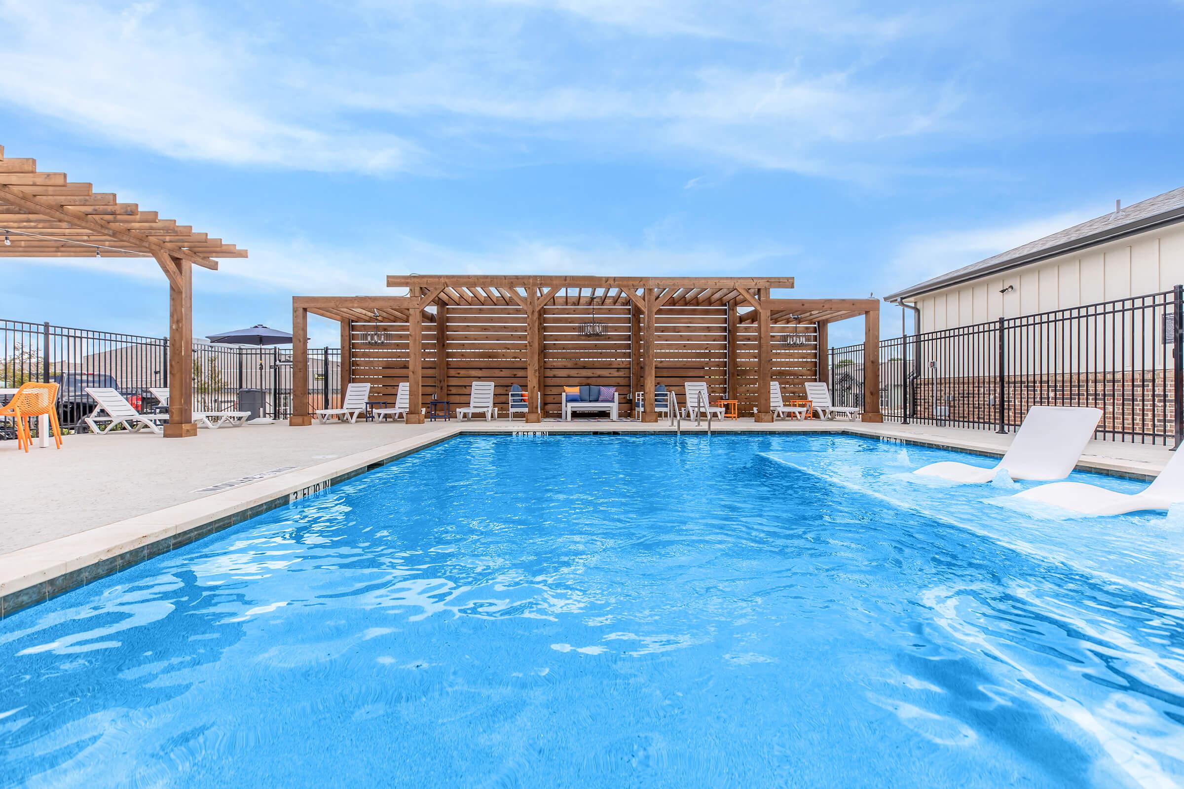 A clear blue swimming pool surrounded by a patio area, featuring lounge chairs and a wooden pergola in the background. The sky is bright with a few clouds, and the setting appears to be a relaxing outdoor space perfect for leisure and recreation.