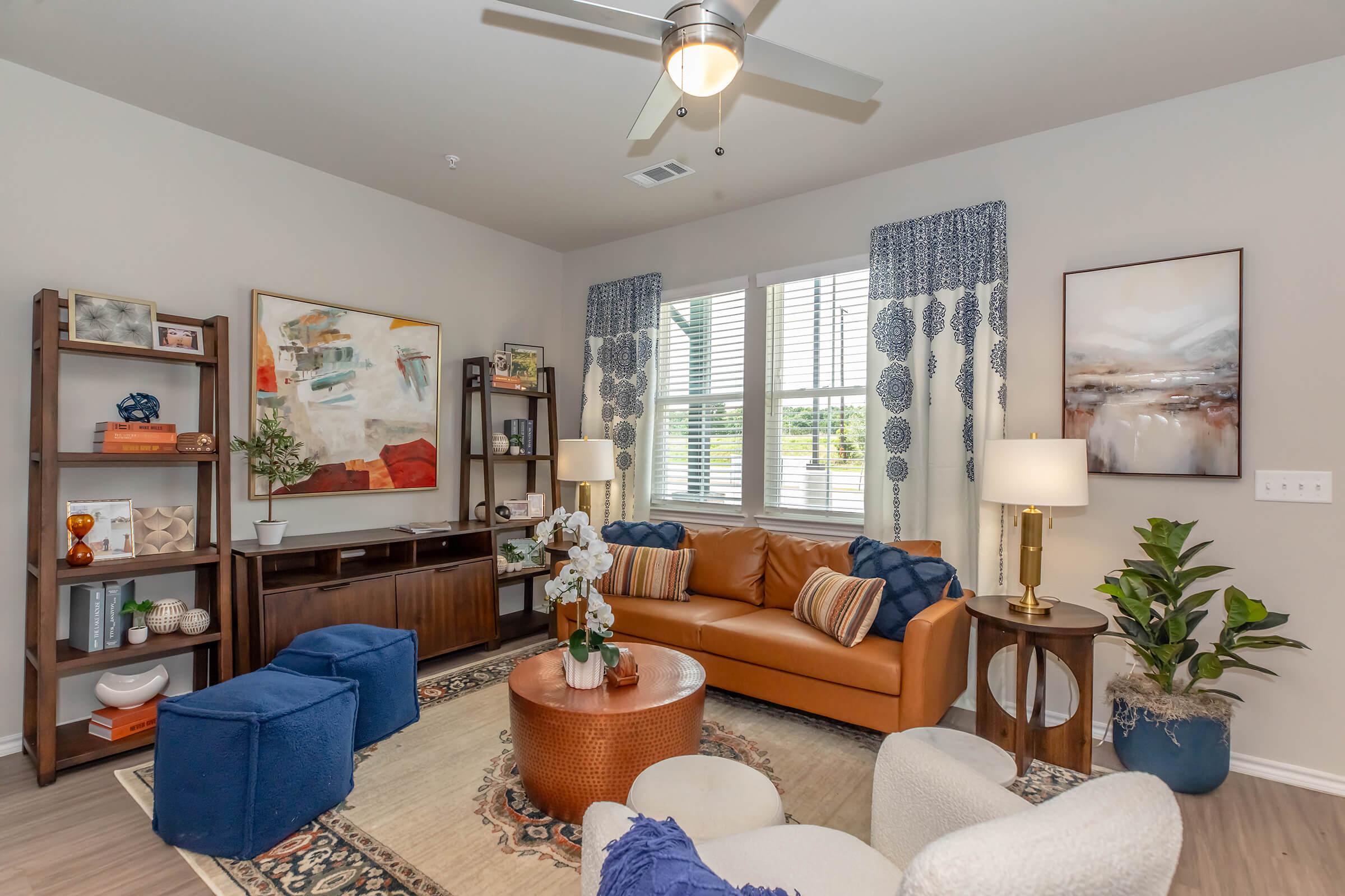 A modern living room with a brown leather sofa, blue ottomans, and a round coffee table. There's a decorative rug on the floor, plants, and a bookshelf with stylish decor. Large windows with blue curtains let in natural light, creating a cozy and inviting atmosphere. Artwork hangs on the walls.