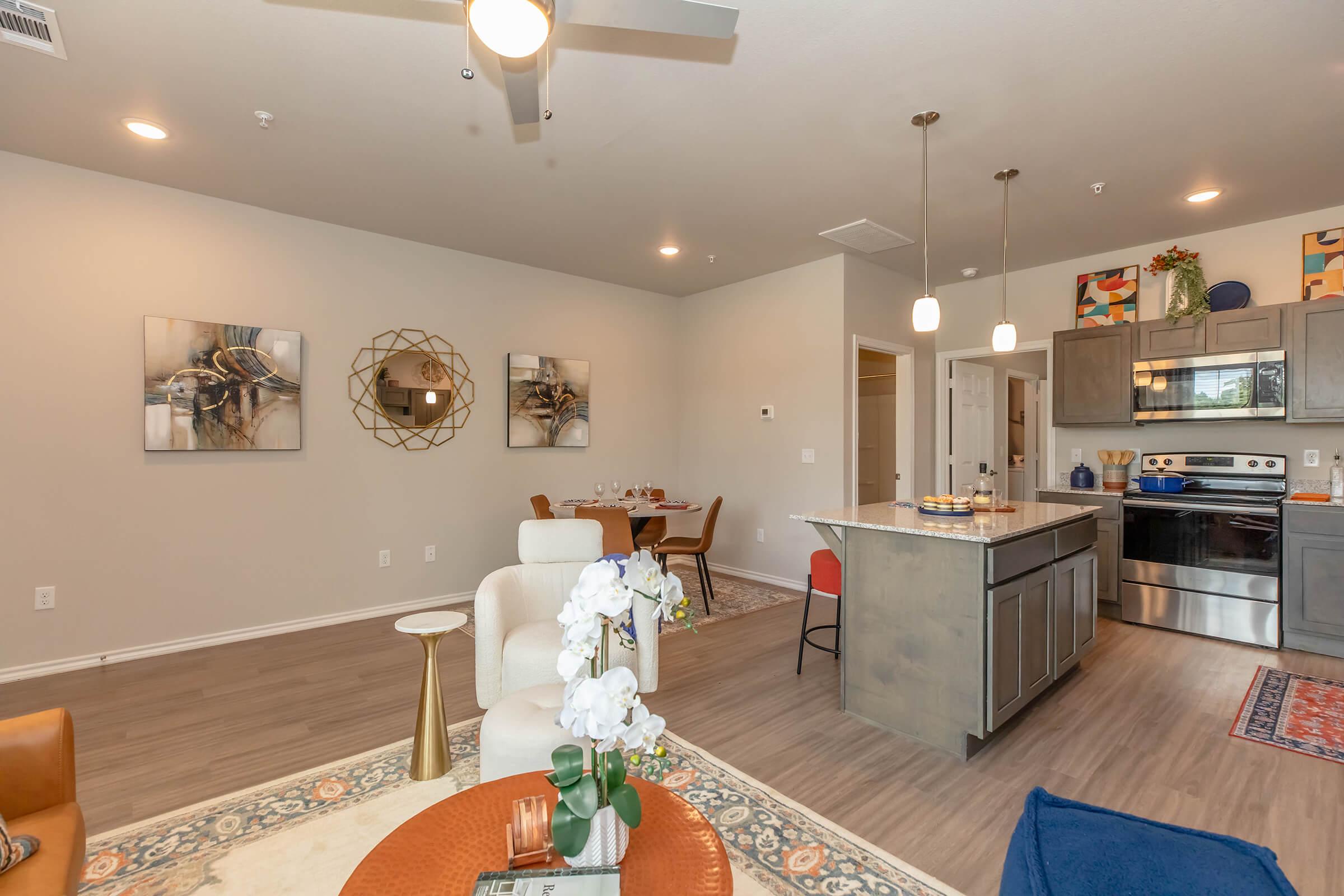 A modern living space featuring an open floor plan with a kitchen and dining area. The kitchen includes stainless steel appliances and a kitchen island. Complemented by stylish decor, the room has light-colored walls, contemporary artwork, a comfortable chair, and a round dining table in the background.
