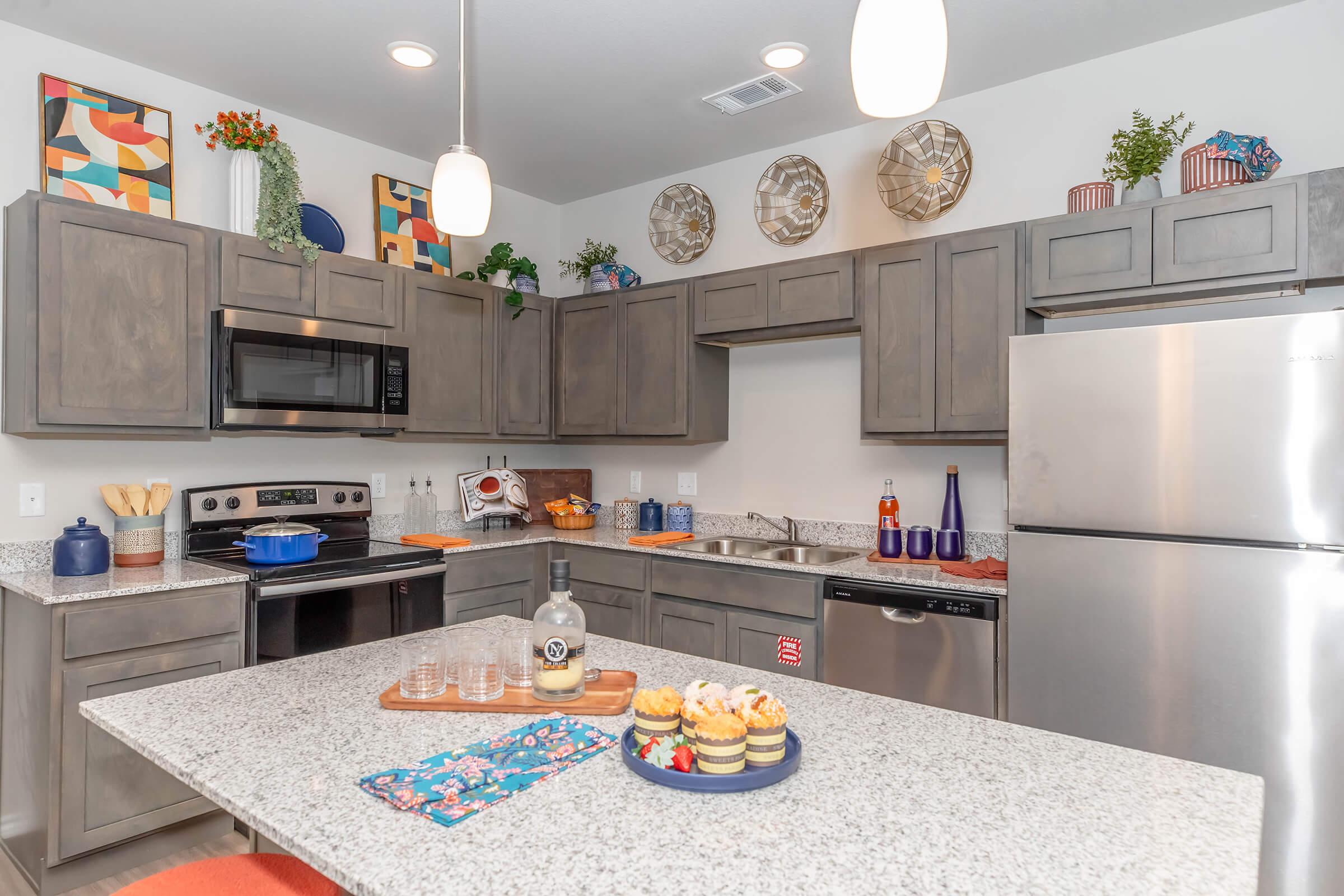 A modern kitchen featuring gray cabinets, stainless steel appliances, and a large island with a granite countertop. Colorful artwork and decorative plants adorn the walls, while a platter of cupcakes and a bottle are placed on the counter, enhancing the inviting atmosphere.