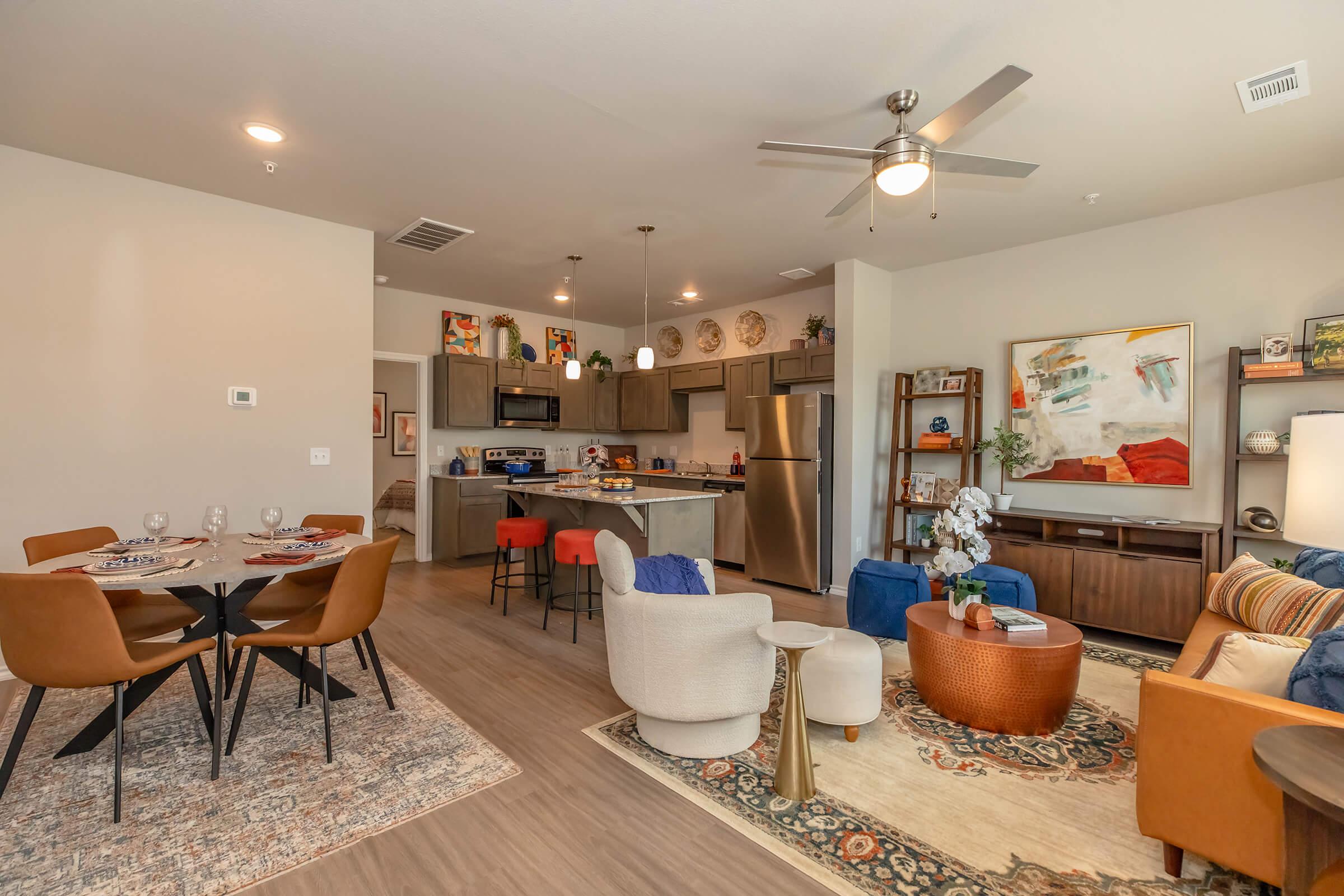 A spacious, modern living area featuring a dining table with four chairs, a cozy seating area with colorful armchairs and a coffee table, and an open kitchen with stainless steel appliances. The decor includes artwork and decorative items, creating a warm and inviting atmosphere.
