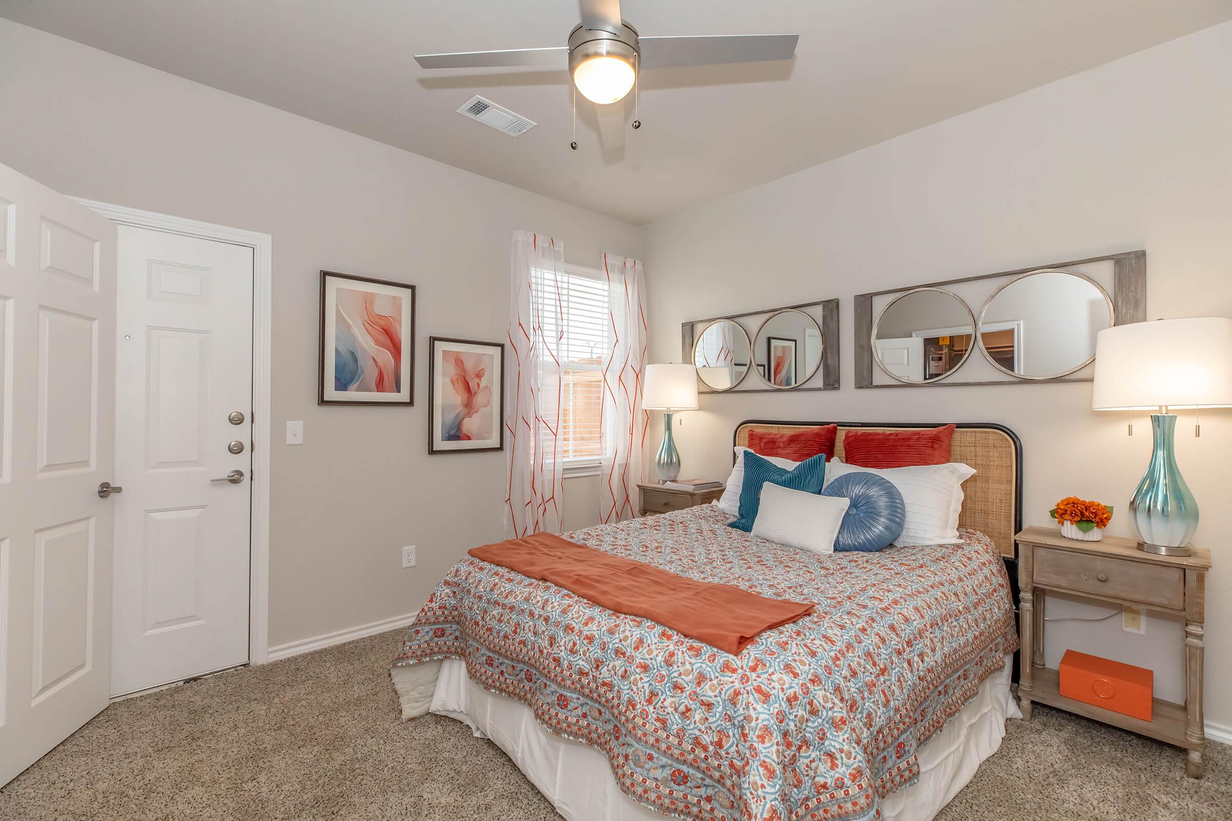 A cozy bedroom featuring a neatly made bed with patterned bedding, decorative pillows, and a plush throw. There are two bedside lamps on wooden nightstands, artwork on the walls, and a ceiling fan. A door leads outside, and windows dressed in light curtains allow natural light to fill the space.