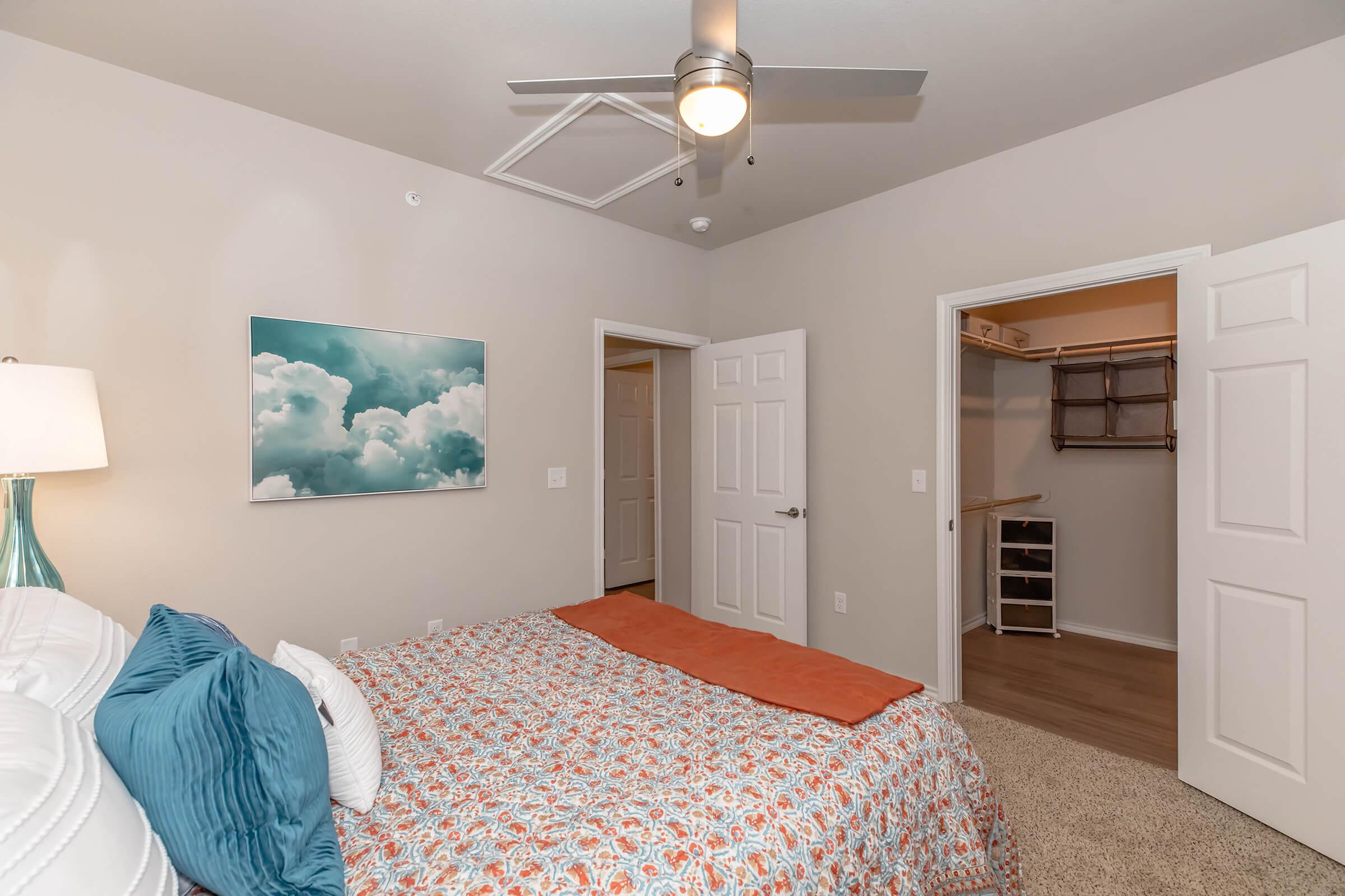 A cozy bedroom featuring a bed with colorful bedding, a bedside lamp, and a wall art piece depicting clouds. The room has a ceiling fan, light-colored walls, and two open doors leading to a closet and another room, enhancing the spacious feel.