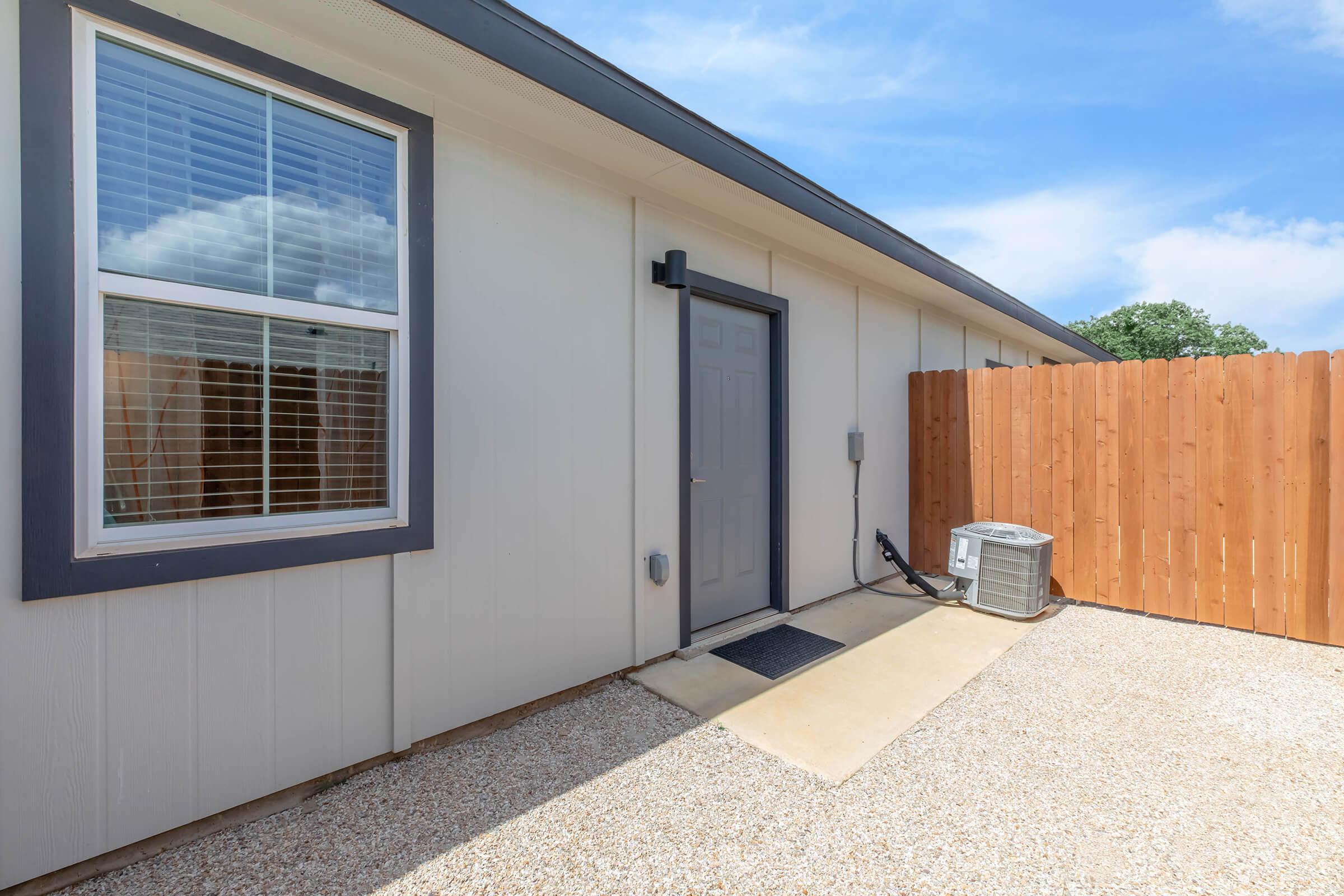 Exterior view of a single-story building with a gray door, a window with blinds, and a gravel path leading to the entrance. There is a wooden fence in the background, and an air conditioning unit is positioned next to the door. The sky is clear with a few clouds.