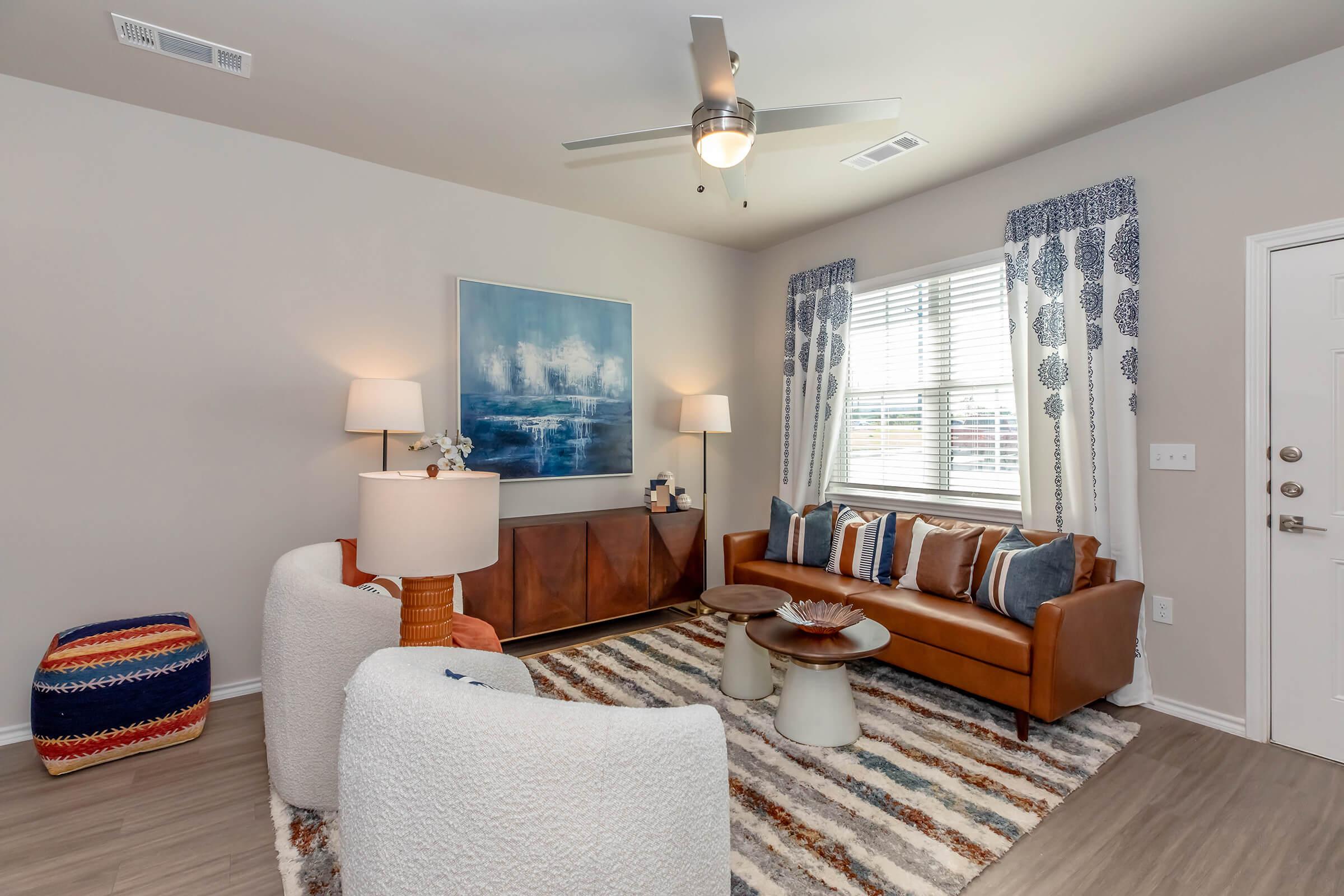 A cozy living room featuring a brown leather sofa with colorful pillows, two white accent chairs, and a striped rug. There's a wooden console table with decorative items, two lamps providing light, and large windows with blue curtains. A ceiling fan adds to the room's comfort.