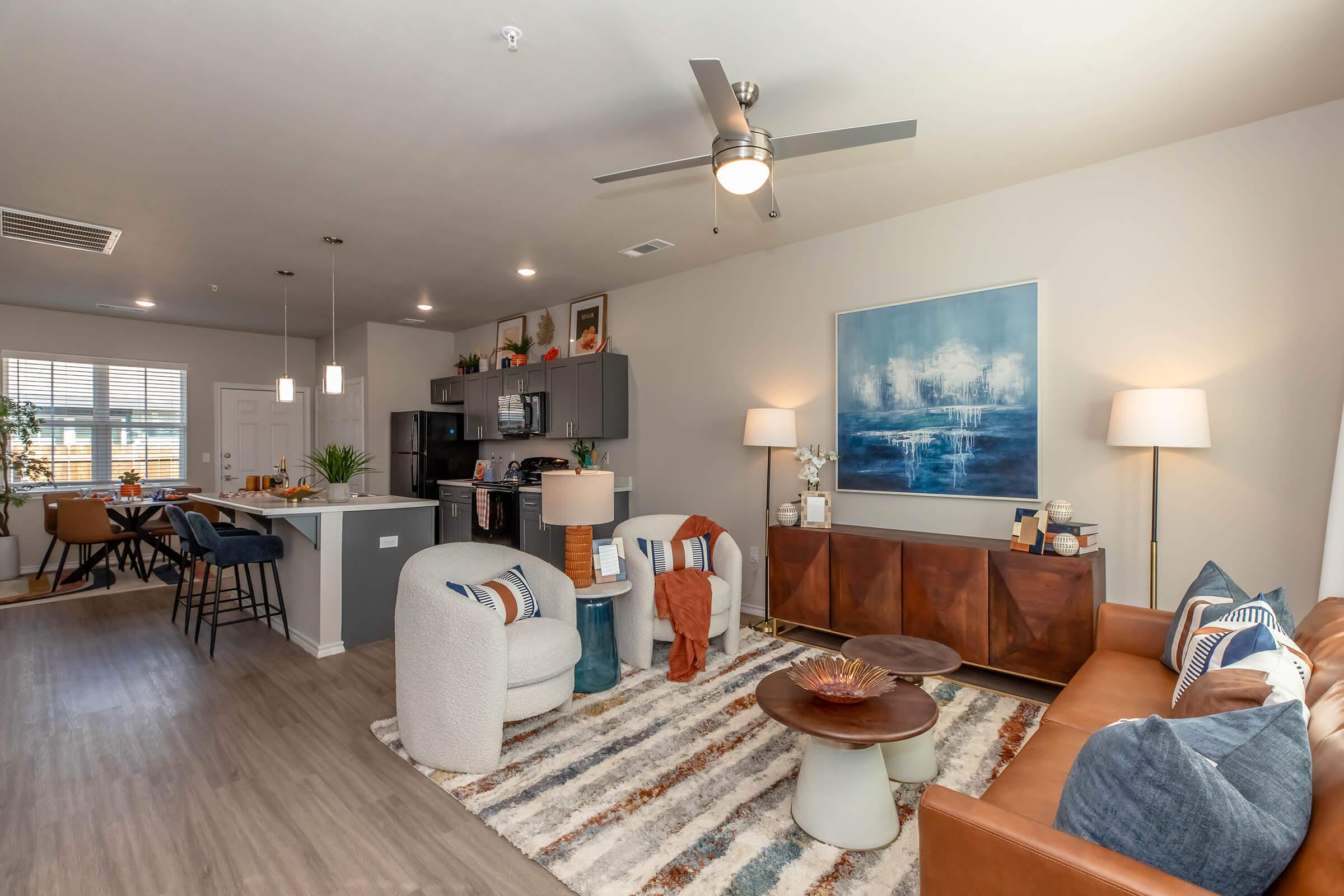 A modern living room featuring a comfortable brown leather sofa and light gray upholstered chairs arranged around a circular coffee table. A colorful area rug covers the floor, and a large abstract painting hangs on the wall. A well-lit kitchen is visible in the background, complete with bar seating and natural light from large windows.