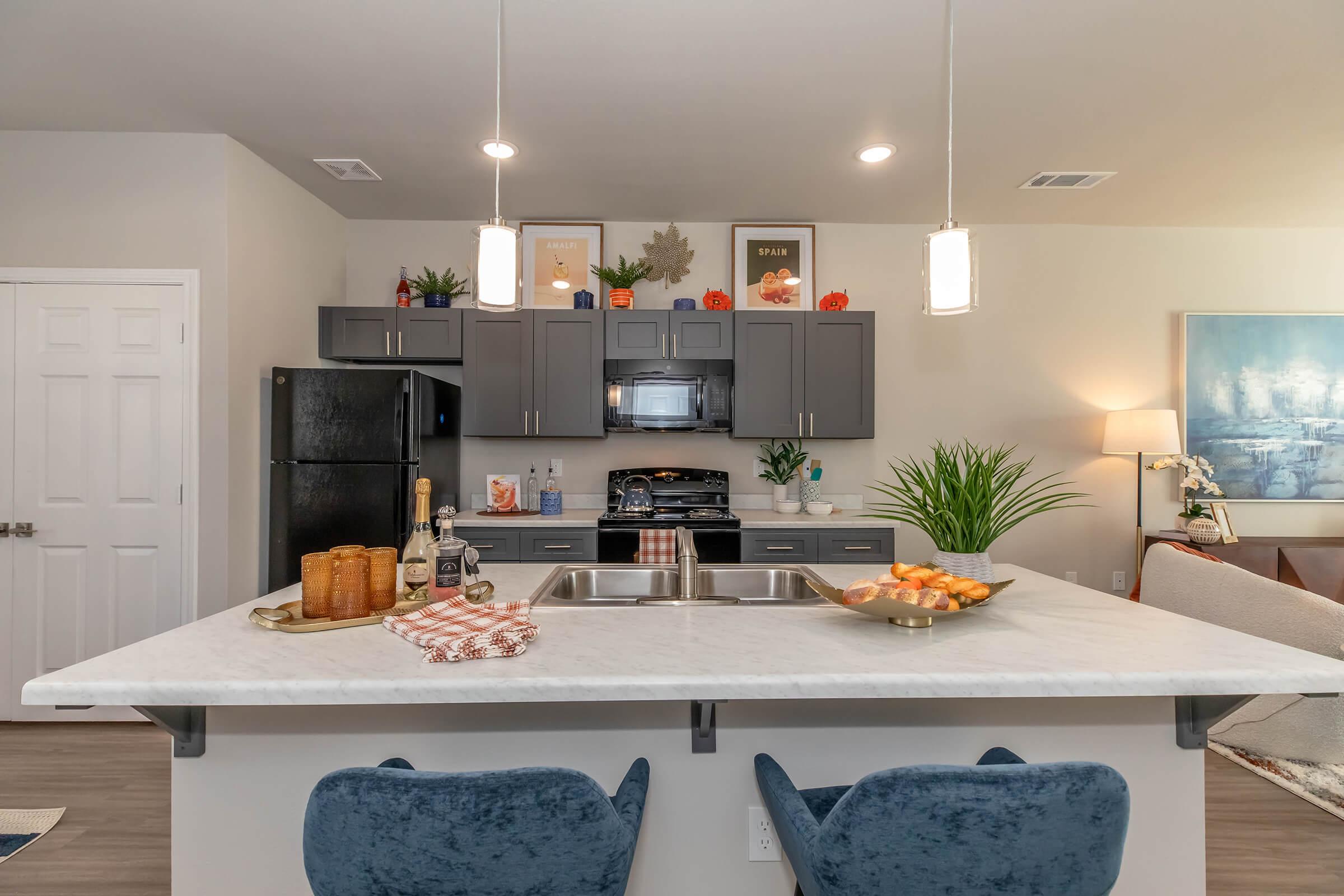 Modern kitchen with dark cabinetry, a white marble island, and two blue chairs. The kitchen is equipped with stainless steel appliances and decorative items, including plants and framed artwork. An inviting atmosphere is created with natural light and stylish decor.
