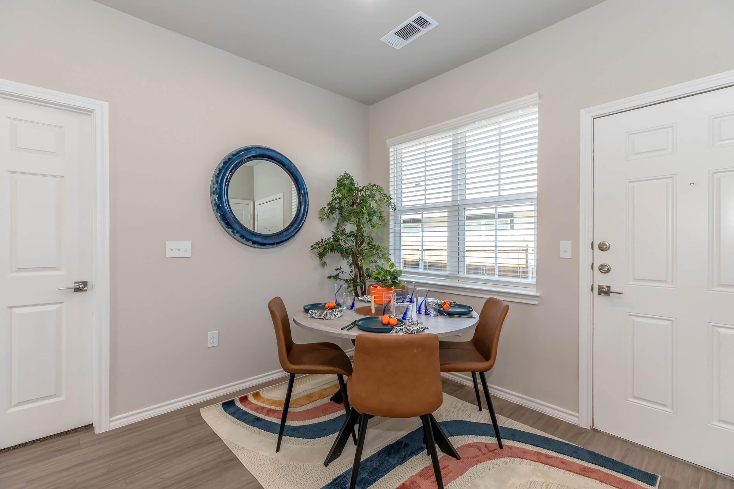 A cozy dining area featuring a round table set for a meal with colorful plates and napkins. There is a decorative mirror on the wall, a small indoor plant on the table, and a large window allowing natural light to brighten the space. The floor is adorned with a stylish area rug.