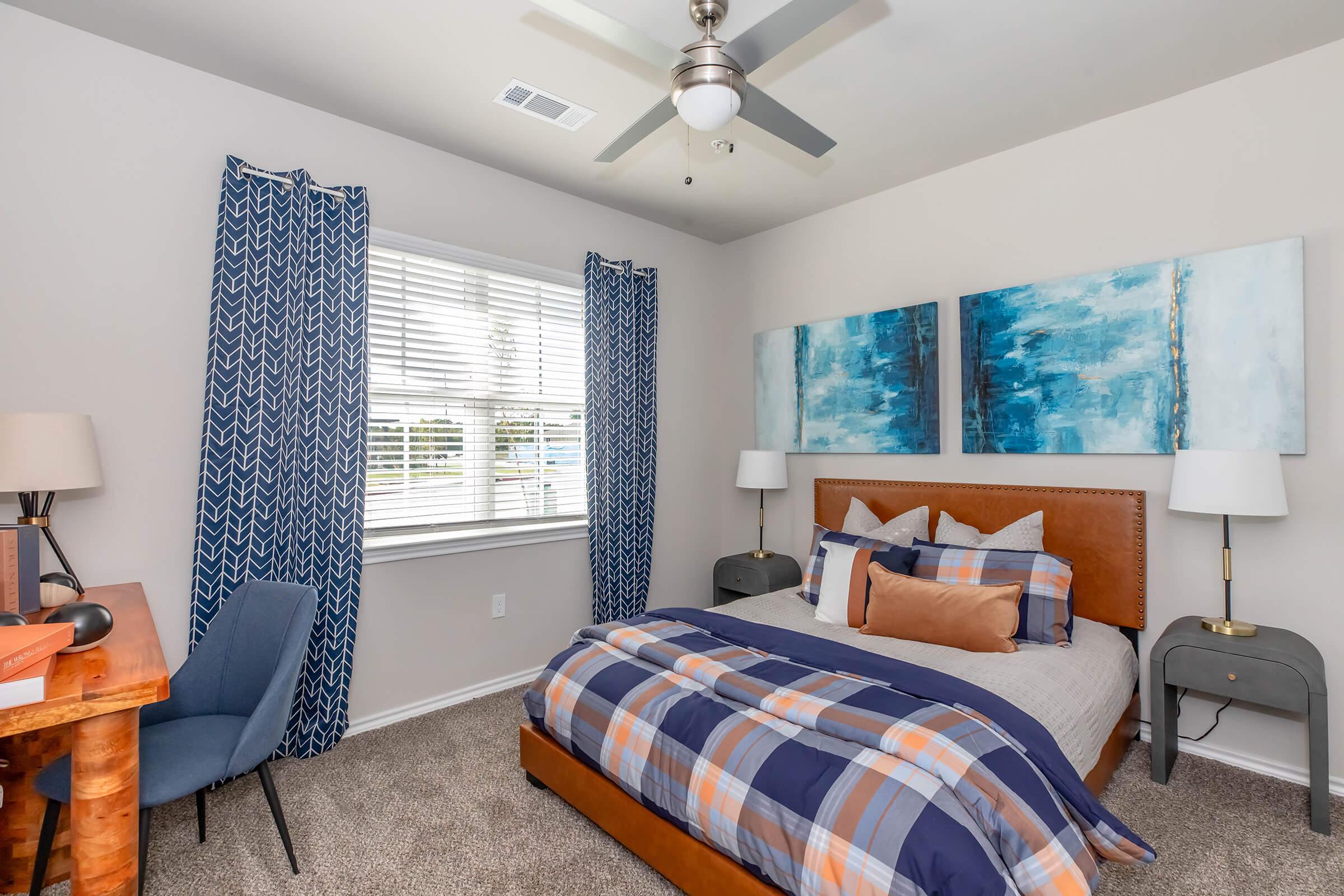 A cozy bedroom featuring a comfortable bed with plaid bedding, decorative pillows, and two large blue abstract paintings above the headboard. The room has light-colored walls, a window with blinds, and blue curtains. A wooden desk and a blue chair are positioned near the window, completing the inviting atmosphere.