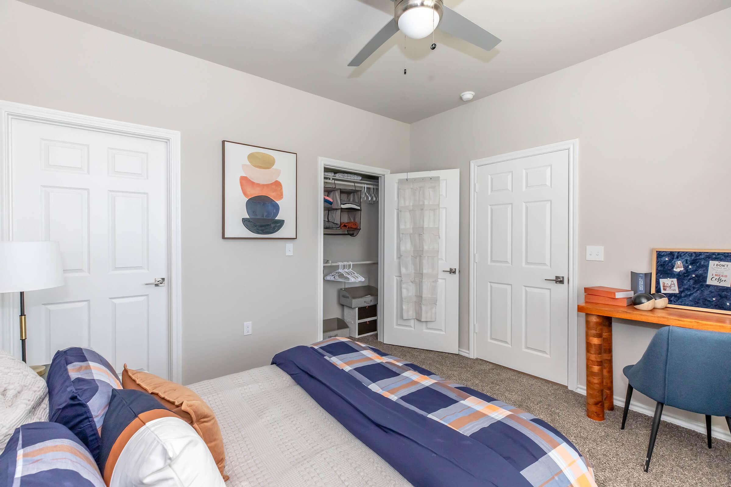 A cozy bedroom featuring a neatly made bed with colorful pillows, a desk with a chair, and a wall art piece. There are two closed doors, one leading to a closet with visible hanging clothes. The room has light-colored walls and a ceiling fan, creating a comfortable atmosphere.