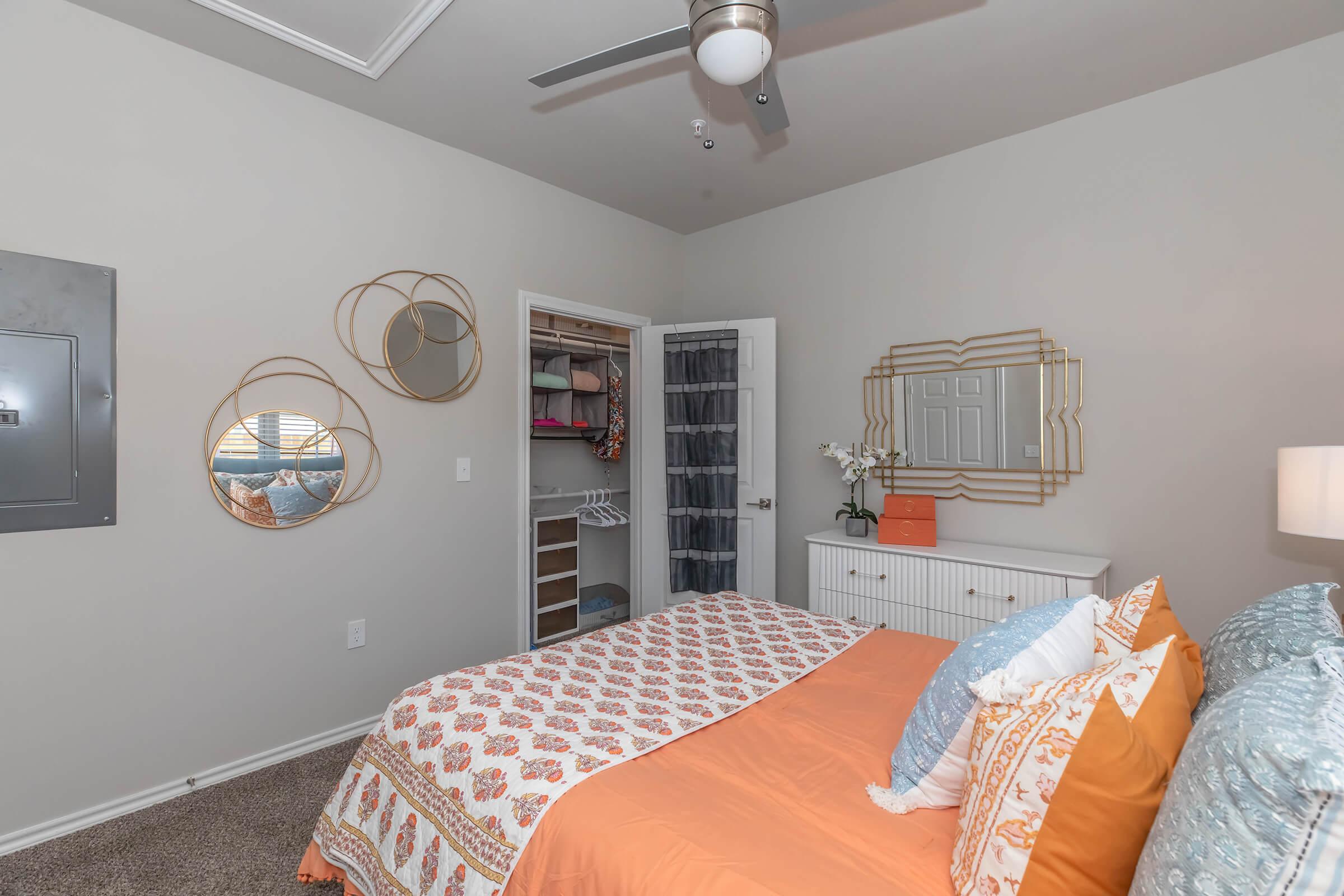 A cozy bedroom featuring a large orange and white patterned bed with decorative pillows. There are two circular mirrors on the wall and a closet with sliding doors. A dresser with a vase is positioned beside the bed, and a ceiling fan provides a modern touch to the space.