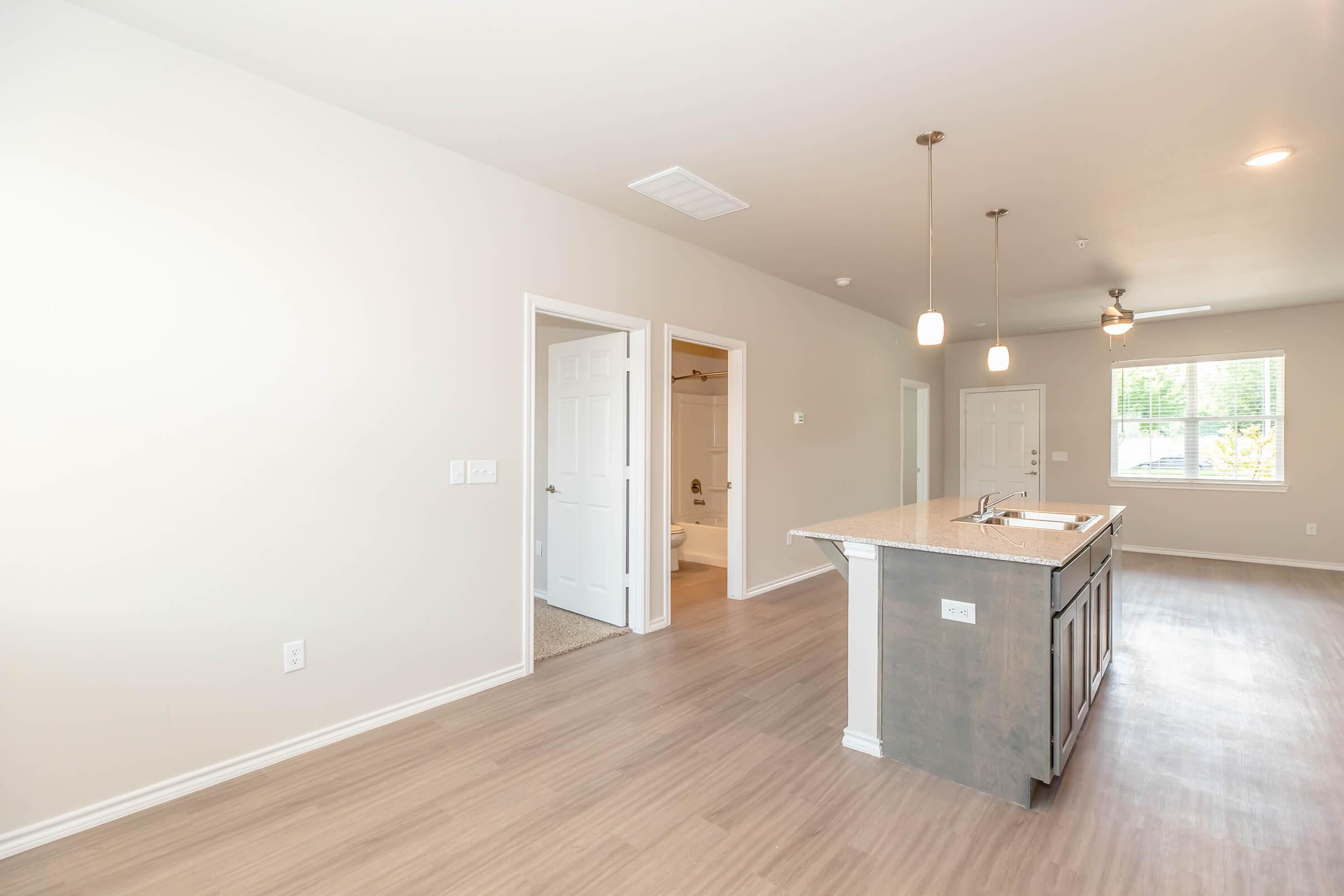 View of a modern, open-concept living space featuring a kitchen island with bar seating, light wood flooring, and neutral-colored walls. Doorways lead to other rooms, including a bathroom visible in the background. Bright natural light comes through a window.