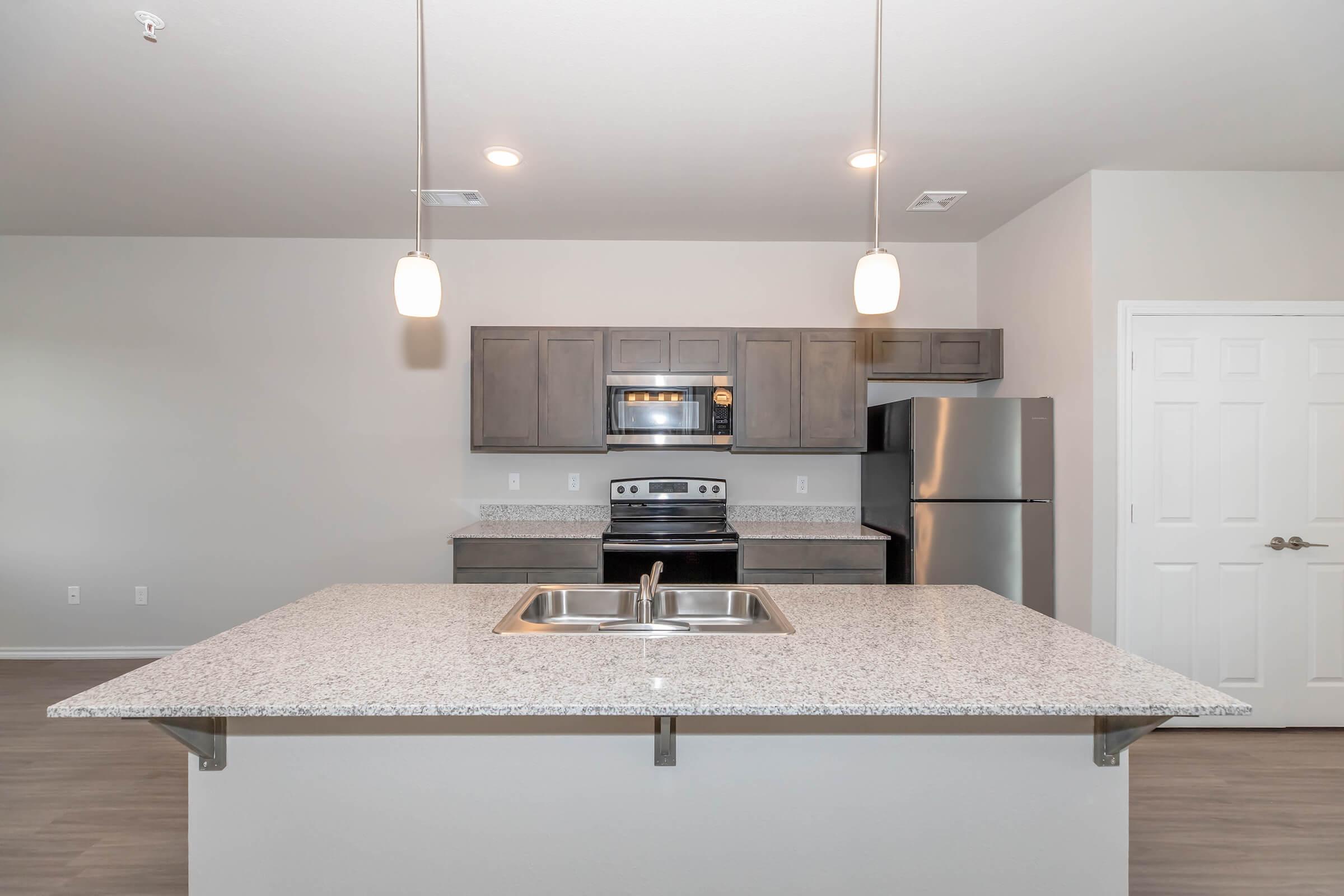 Modern kitchen featuring a central island with a double sink, granite countertops, stainless steel appliances including an oven and refrigerator, gray cabinetry, and pendant lighting. The walls are painted light gray, and the flooring is a wood-like laminate, creating a clean and contemporary look.