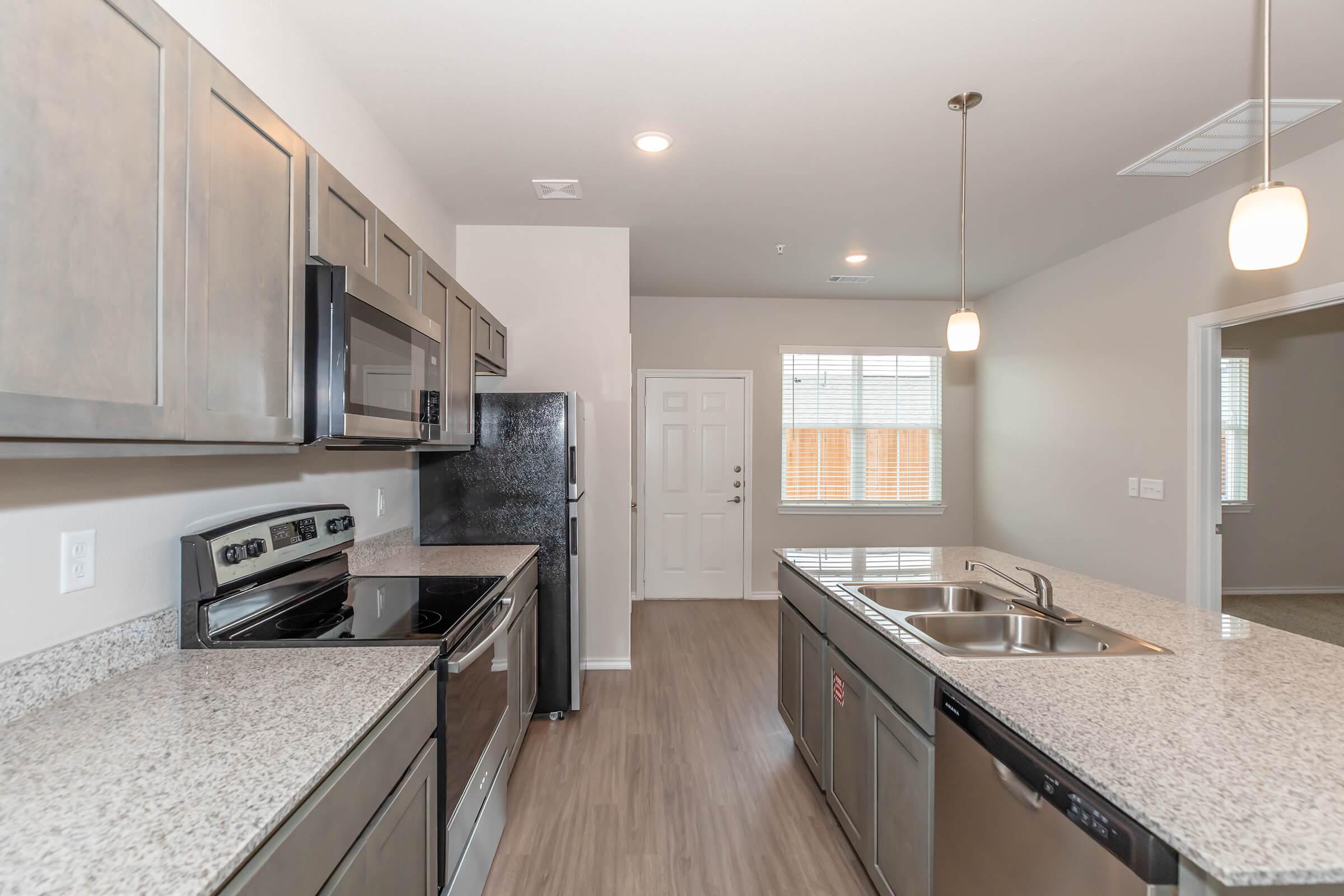 Modern kitchen featuring stainless steel appliances, dark wood cabinets, and granite countertops. A central island with a double sink separates the cooking area from the living space. Natural light enters through a window, and there is a door leading outside, enhancing the open and airy feel of the room.