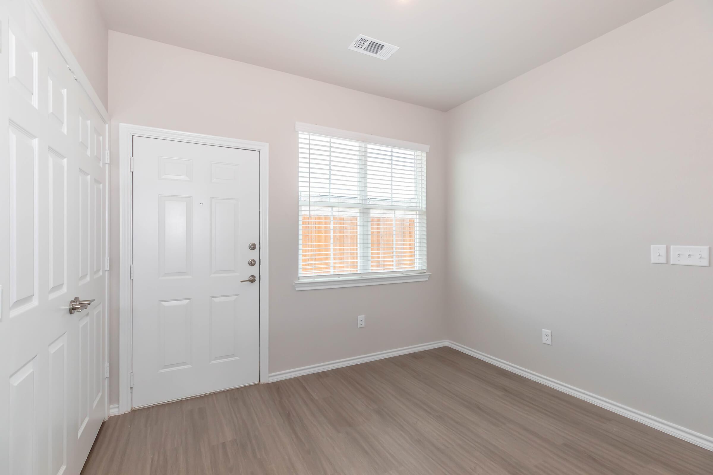 A small, empty room featuring light-colored walls and a wooden floor. There is a white door on the left and a window with blinds providing natural light. The room appears bright and minimalistic, suitable for use as an office or small living space.