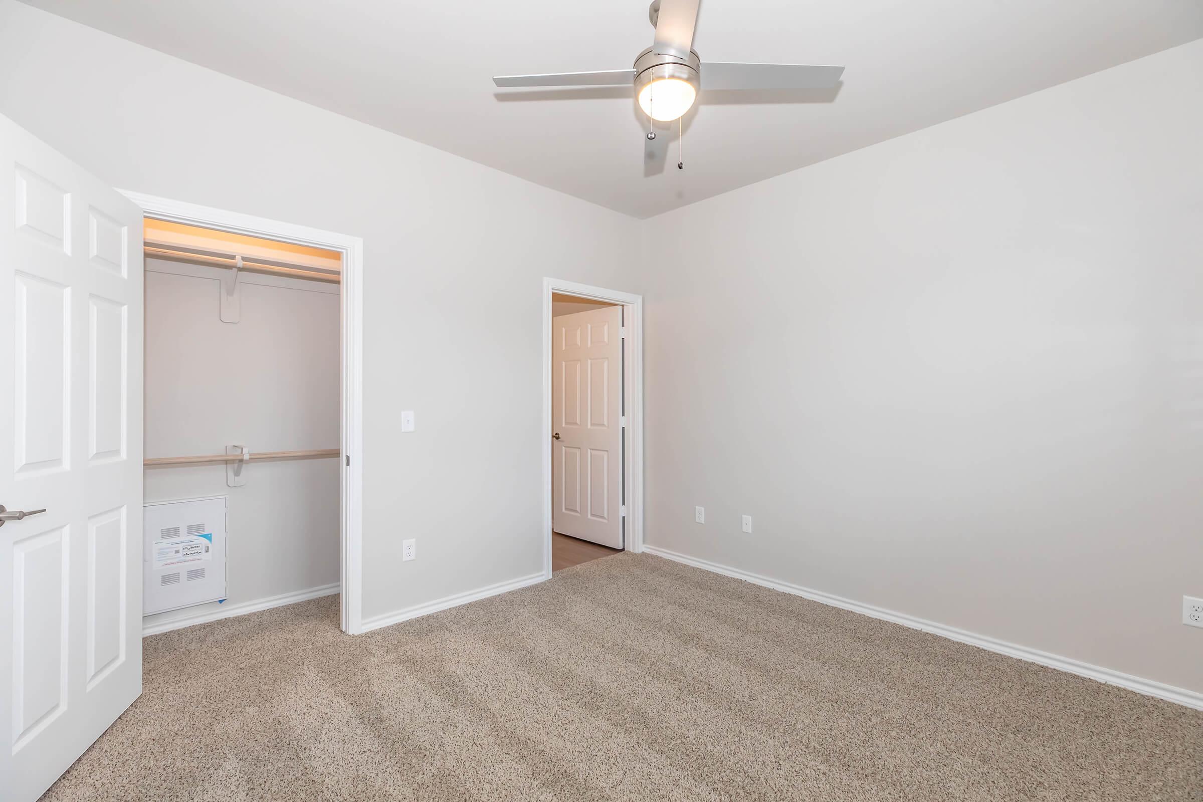 A well-lit, empty bedroom featuring light-colored walls, beige carpeted flooring, and a ceiling fan. Open doors lead to a closet and an adjacent room, creating a spacious feel. The room is unfurnished, providing an opportunity for personalization.