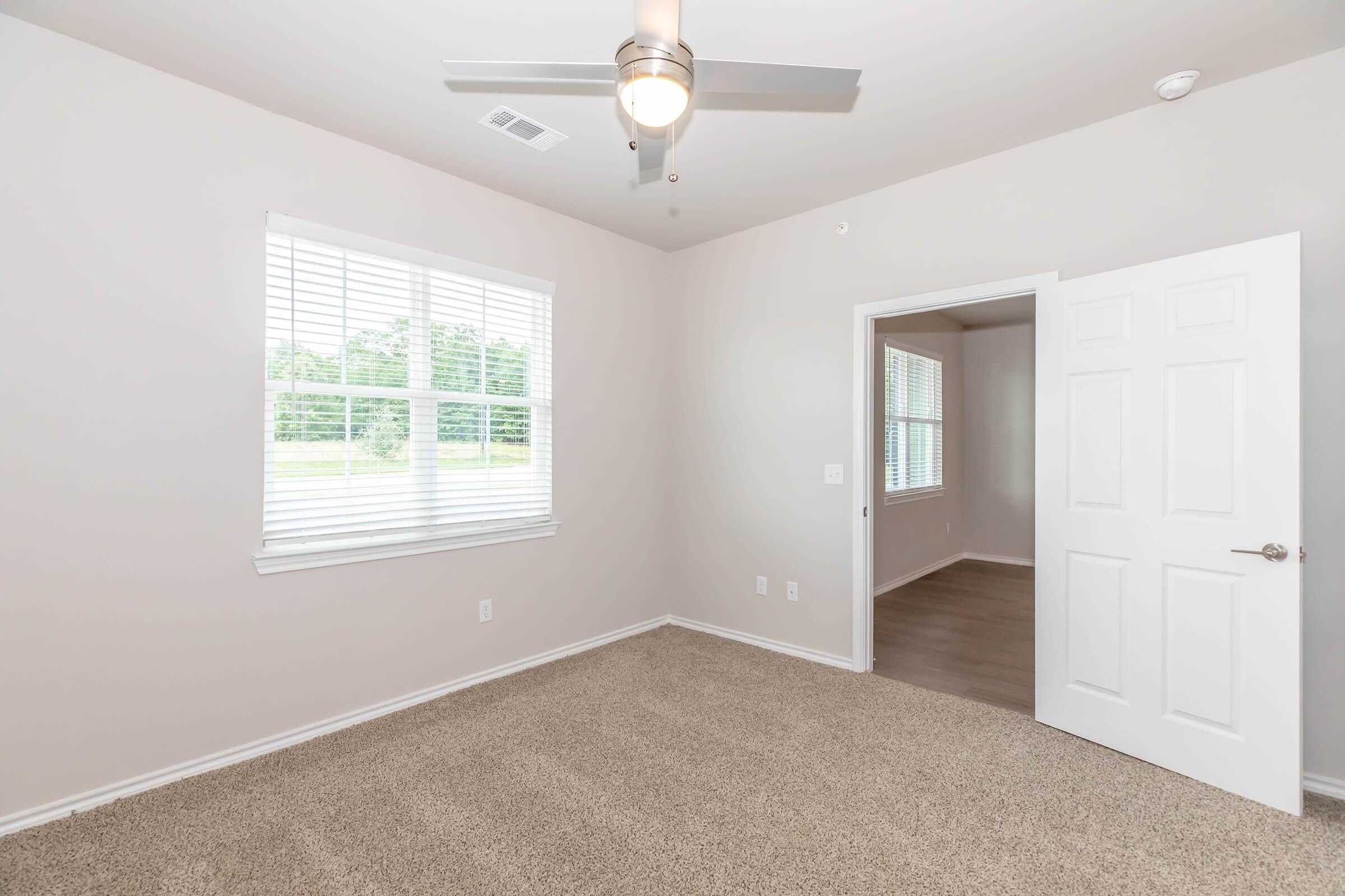 A well-lit, empty room with beige carpeting, a ceiling fan, and white walls. One window with blinds offers natural light, while a door open to an adjacent room is visible. The space is perfect for a bedroom or office, creating a cozy atmosphere with its neutral color palette.