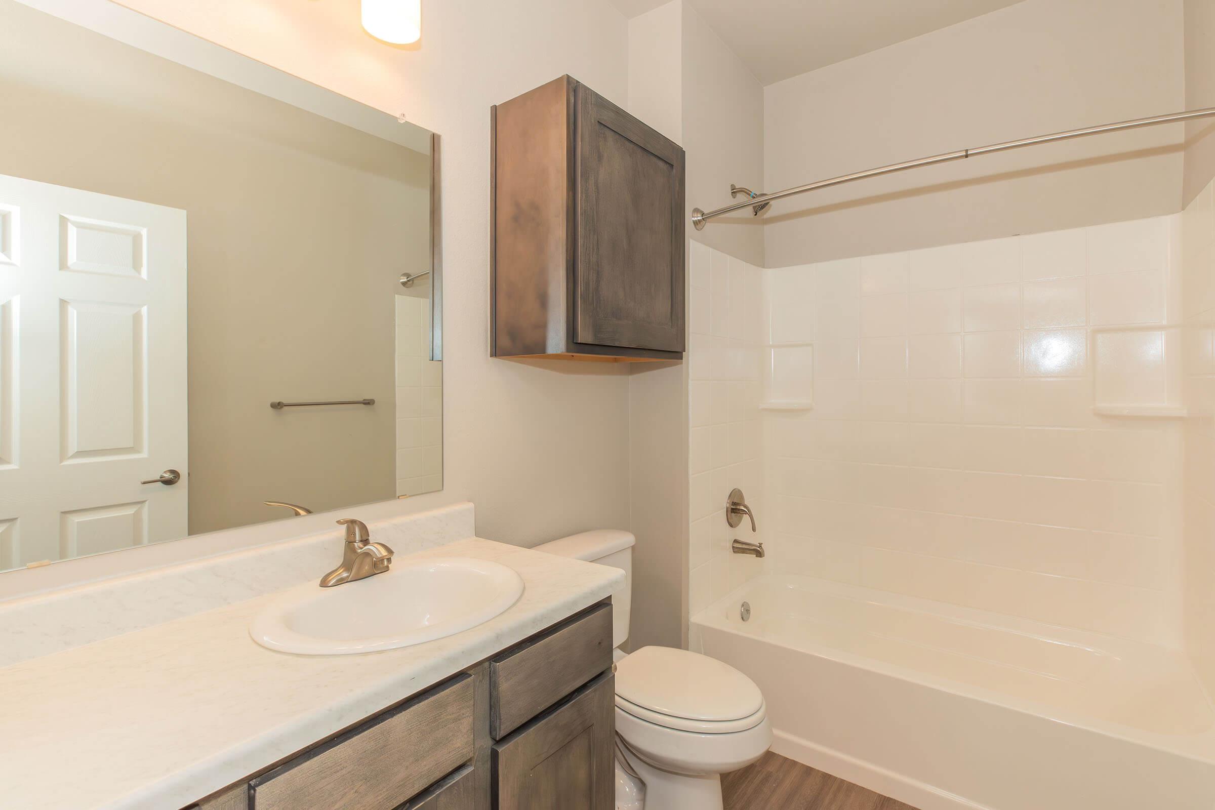 A modern bathroom featuring a white bathtub and shower, a single sink with a marble countertop, and a dark wooden cabinet above the sink. The walls are light-colored, and there is a mirror above the sink. A towel rod is visible on one wall, and the flooring is a wood-like laminate.