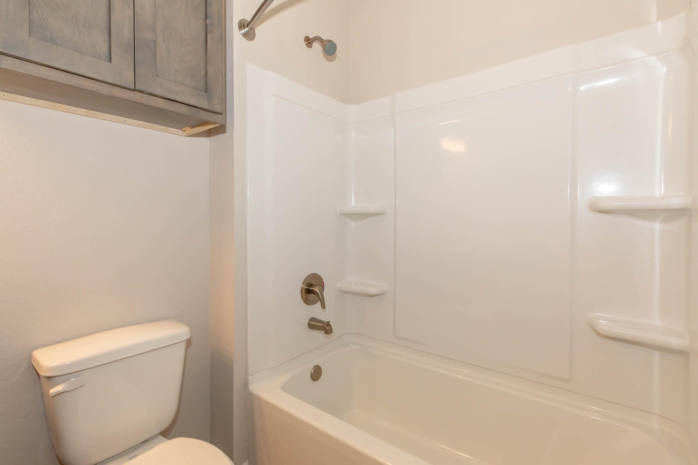 A clean, modern bathroom featuring a white bathtub with shower fixtures, a beige wall, and gray cabinetry above. The room includes a white toilet positioned beside the bathtub. The overall aesthetic is bright and uncluttered, providing a fresh and inviting atmosphere.