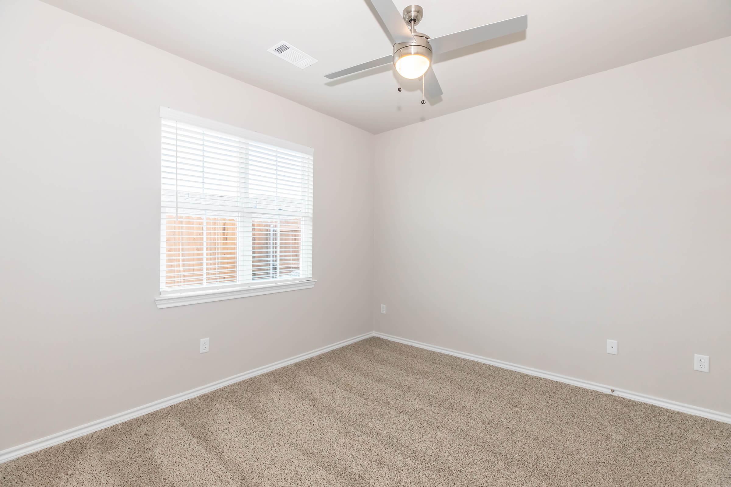 Empty room with light beige walls and carpet. A window with white blinds allows natural light to enter. A modern ceiling fan is installed in the center of the ceiling. The room has a clean, minimalistic appearance with no furniture or decorations.