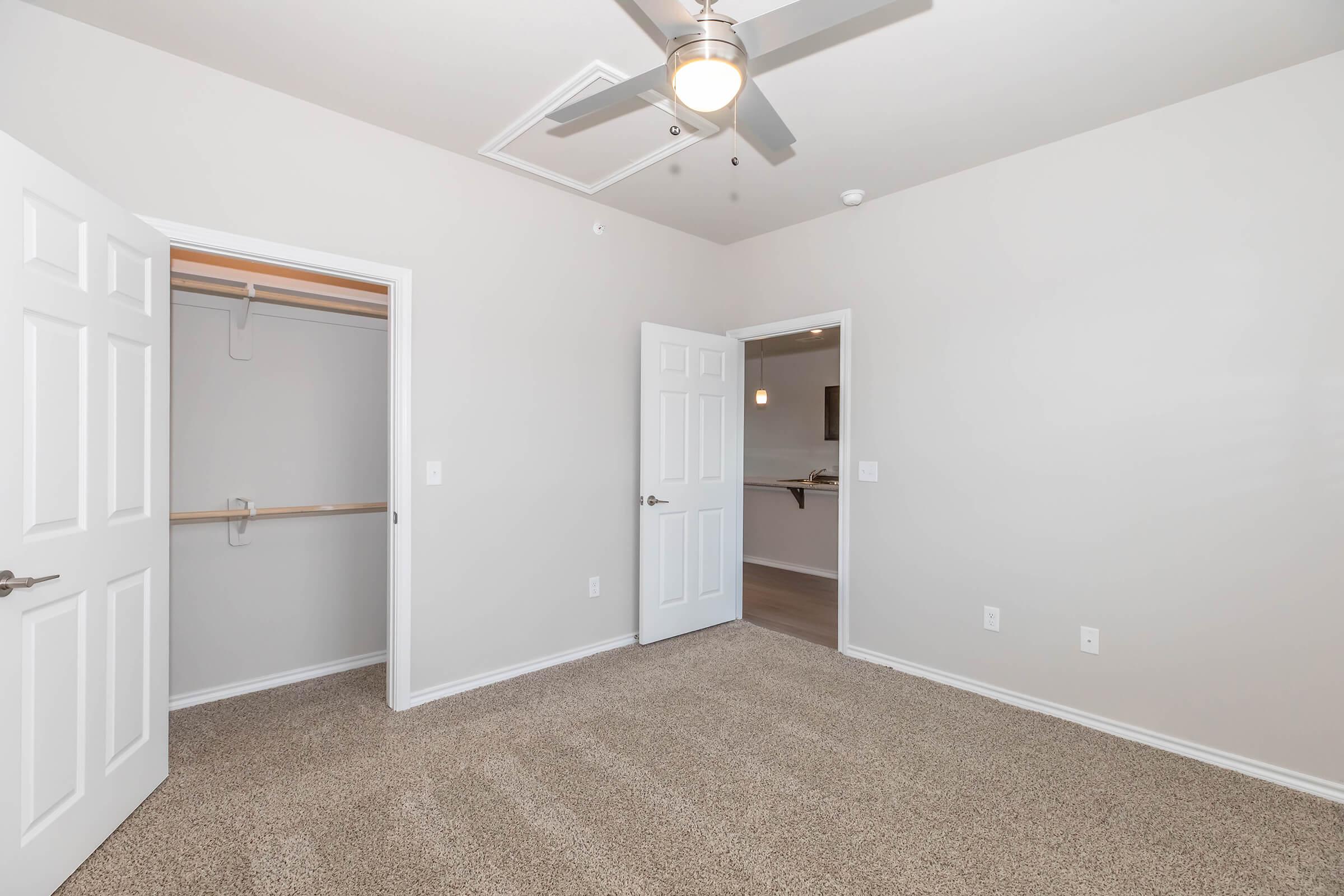 A spacious, empty room featuring light-colored walls and plush carpeting. There is a ceiling fan, a closet with sliding doors, and an adjoining door leading to another area. The room is well-lit with natural light and has a neutral aesthetic, making it versatile for various uses.