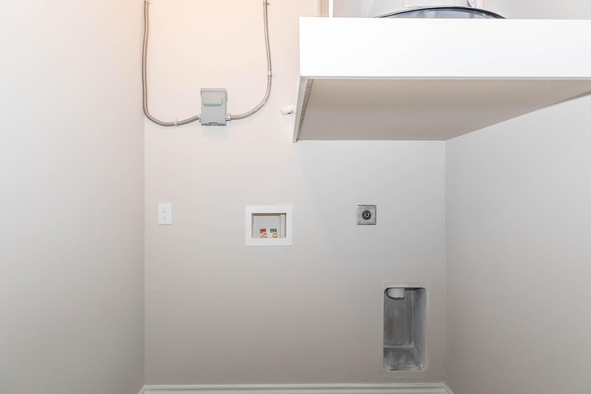 A clean, empty laundry room with light-colored walls. There is a small electrical panel on the wall, a recessed area with pipes, and a shelf above, but no appliances or furniture present. The floor is bare and the overall space looks well-maintained and organized.