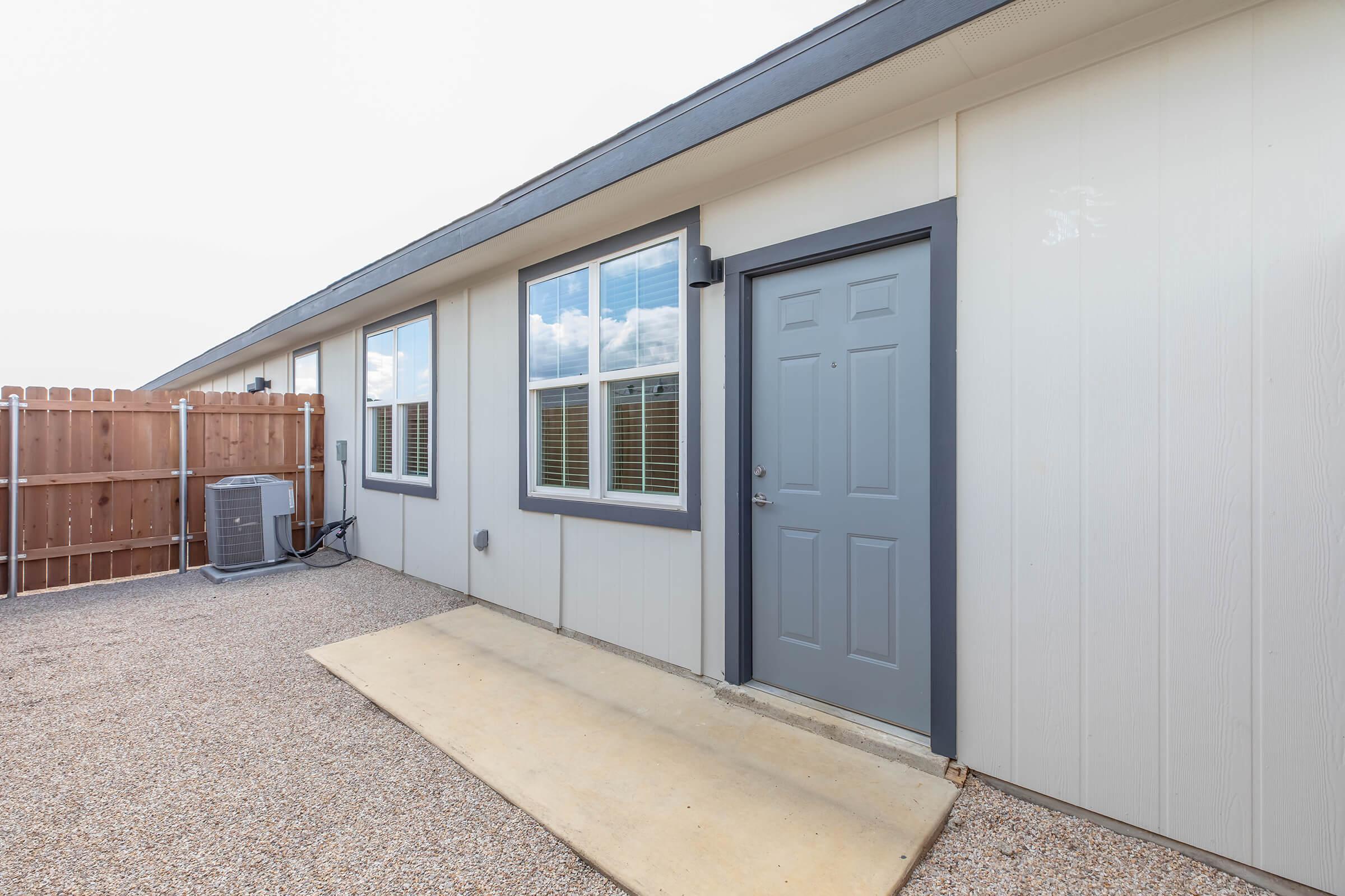 A view of the exterior of a modern home showing a gray door, windows with white frames, and a small concrete porch area. The side of the house features a fenced area and an air conditioning unit. The landscape is gravel with no visible vegetation.
