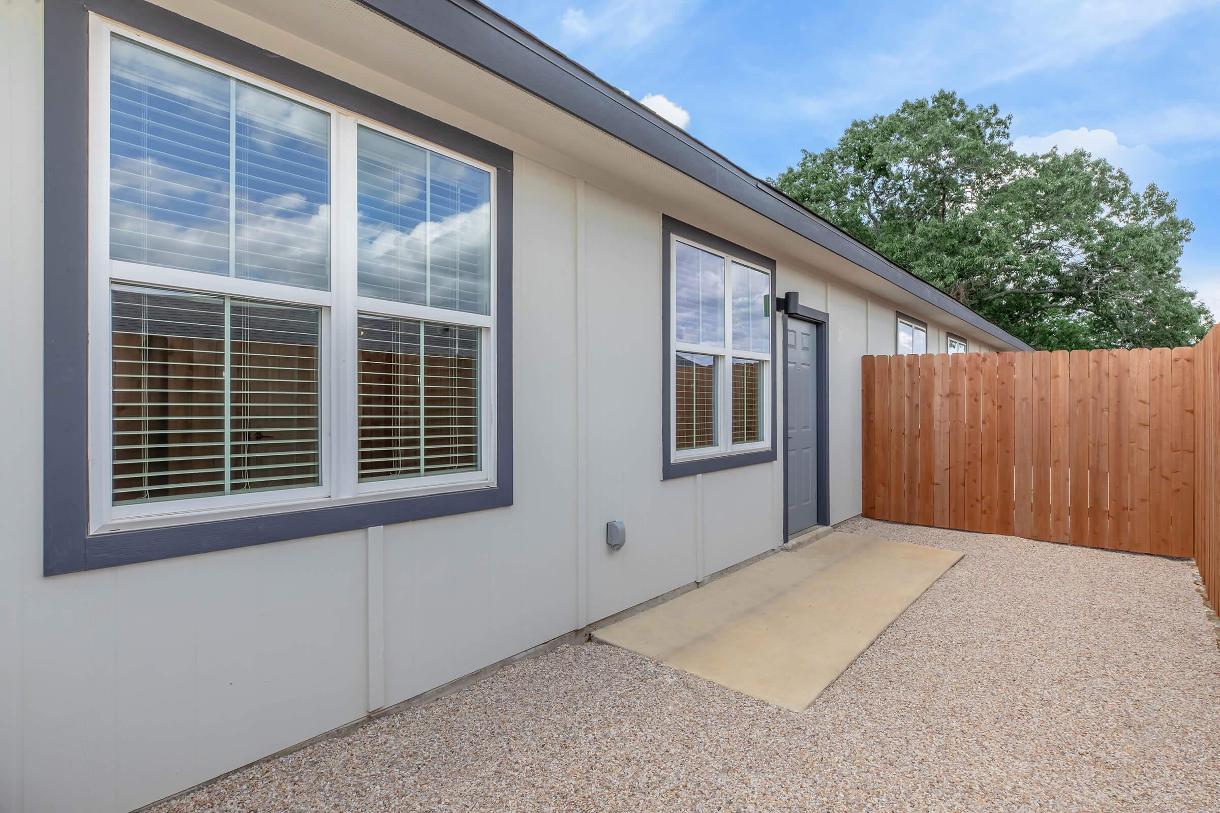 Exterior view of a modern single-story building featuring large windows with blinds, a gray door, and a small gravel area in front. A wooden fence runs alongside the property, providing privacy. The sky is partly cloudy, adding to the tranquil atmosphere.
