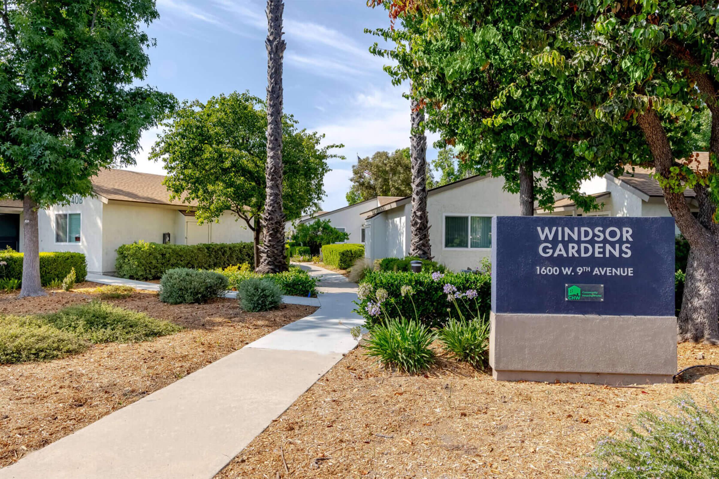 Pathway leading through landscaped gardens with palm trees and shrubs, leading to a building complex. A sign on the right displays "Windsor Gardens 1600 W 9th Avenue." The scene is bright and inviting, depicting a serene residential environment.