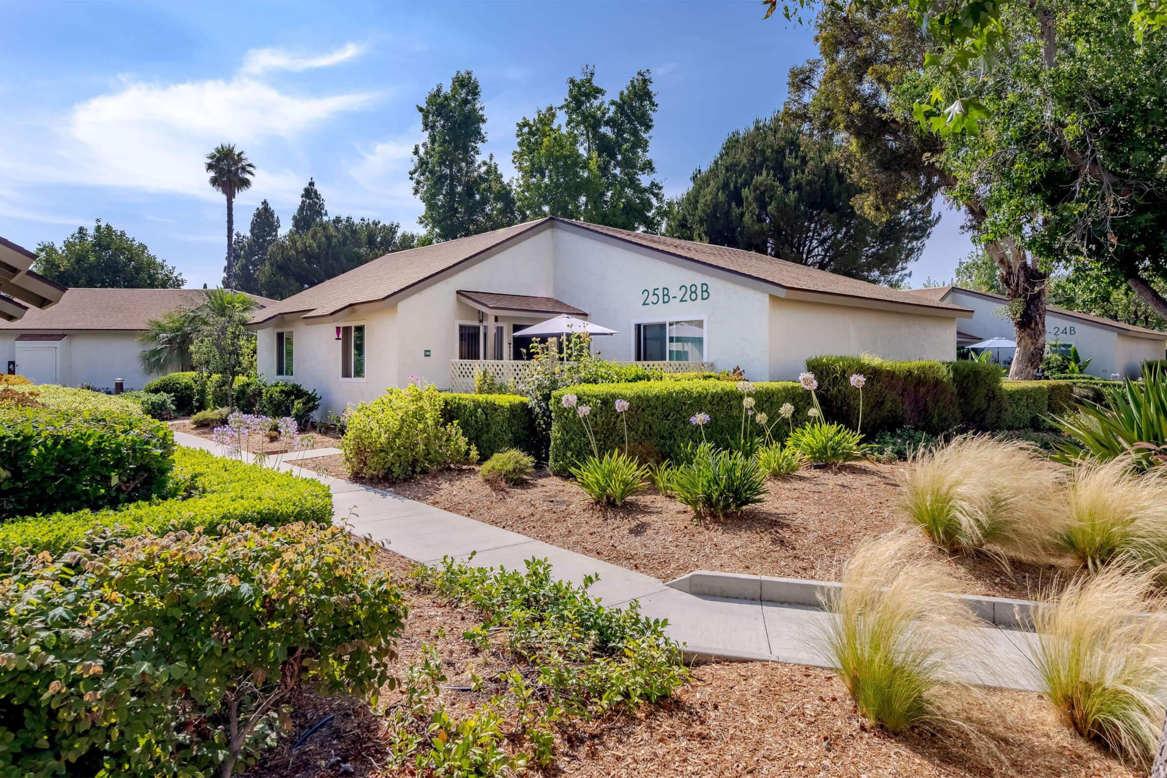 A charming single-story house with a light-colored exterior, surrounded by lush greenery and colorful flowers. A well-maintained pathway leads to the entrance, with a few palm trees in the background. The sky is clear and blue, creating a pleasant atmosphere.