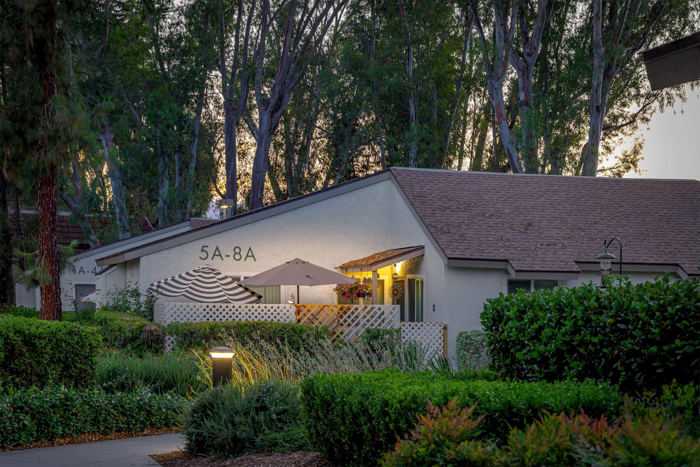 A cozy residential building at dusk, featuring a patio with umbrellas and outdoor furniture. Lush greenery and well-maintained landscaping surround the area, with soft lighting illuminating the path. The scene reflects a peaceful and inviting atmosphere.