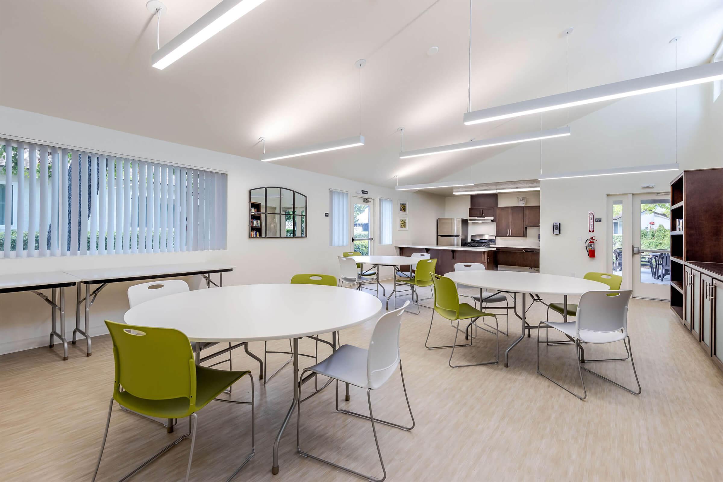 Interior view of a modern dining area with multiple round tables and chairs. The space features large windows with vertical blinds, bright lighting, and a kitchen area visible in the background. The walls are light-colored, and the floor has a wood-like finish, creating a welcoming atmosphere.