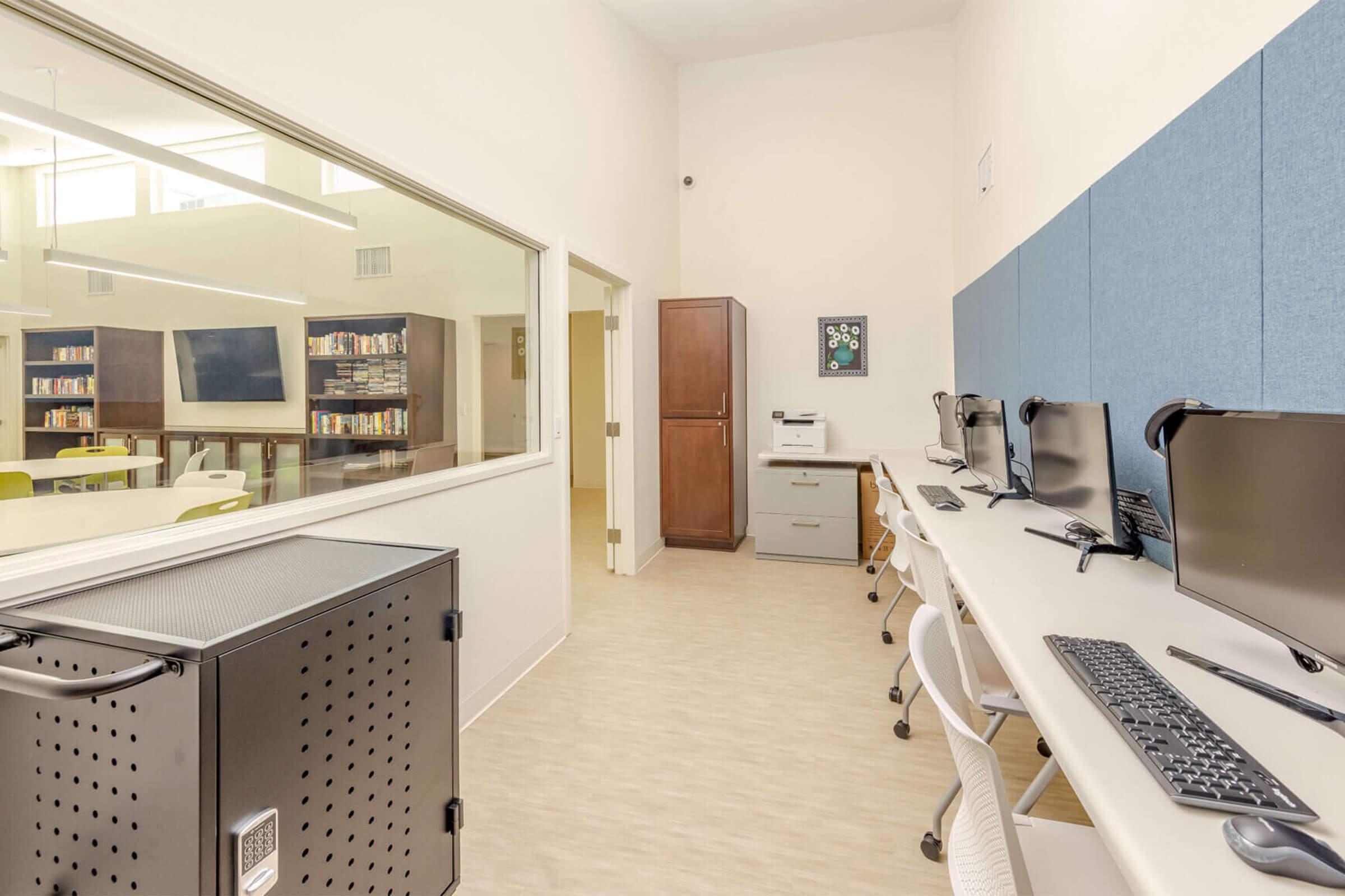 A modern office space featuring a row of computers with ergonomic chairs, light-colored walls, and a large window providing natural light. In the background, shelves filled with books are visible, along with a flat-screen TV and a printer. The room has a clean, organized appearance.