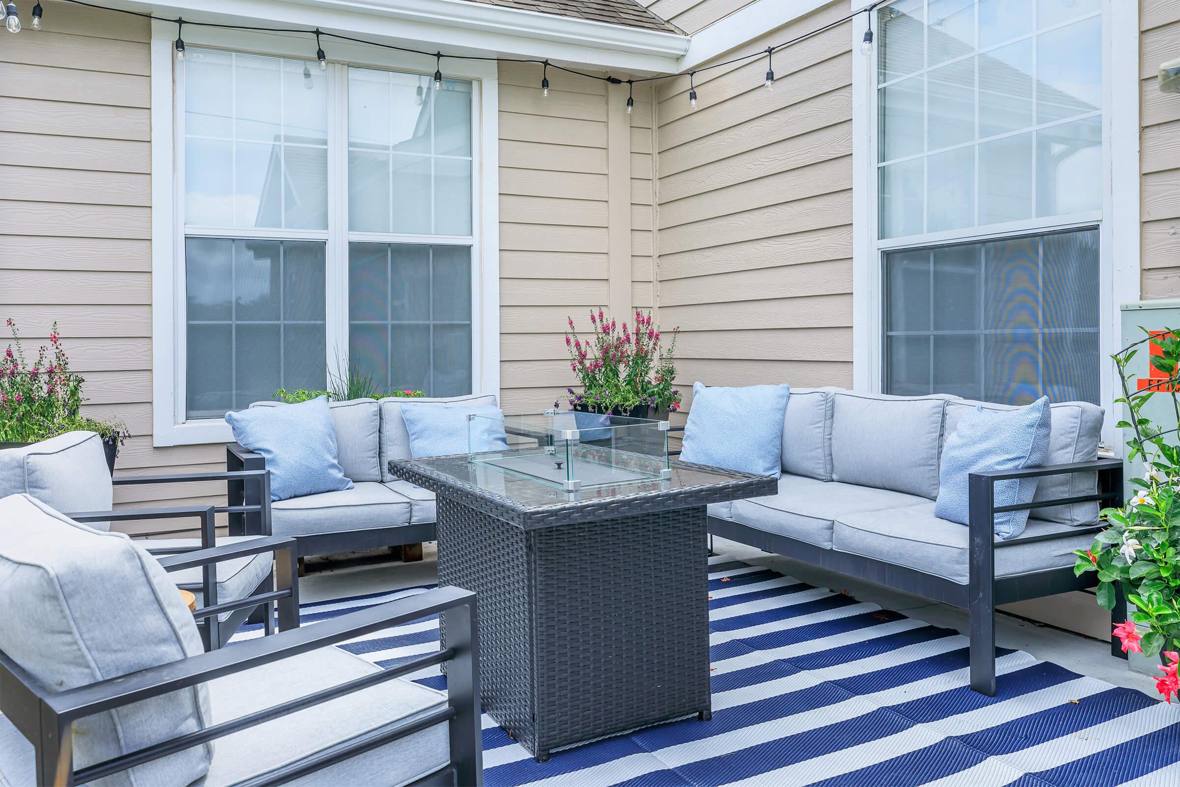 A cozy outdoor patio setting featuring a seating arrangement with light blue cushions and a glass-top coffee table. The area is adorned with potted plants and greenery, and a striped blue and white rug covers the floor, creating a welcoming atmosphere for relaxation or socializing.