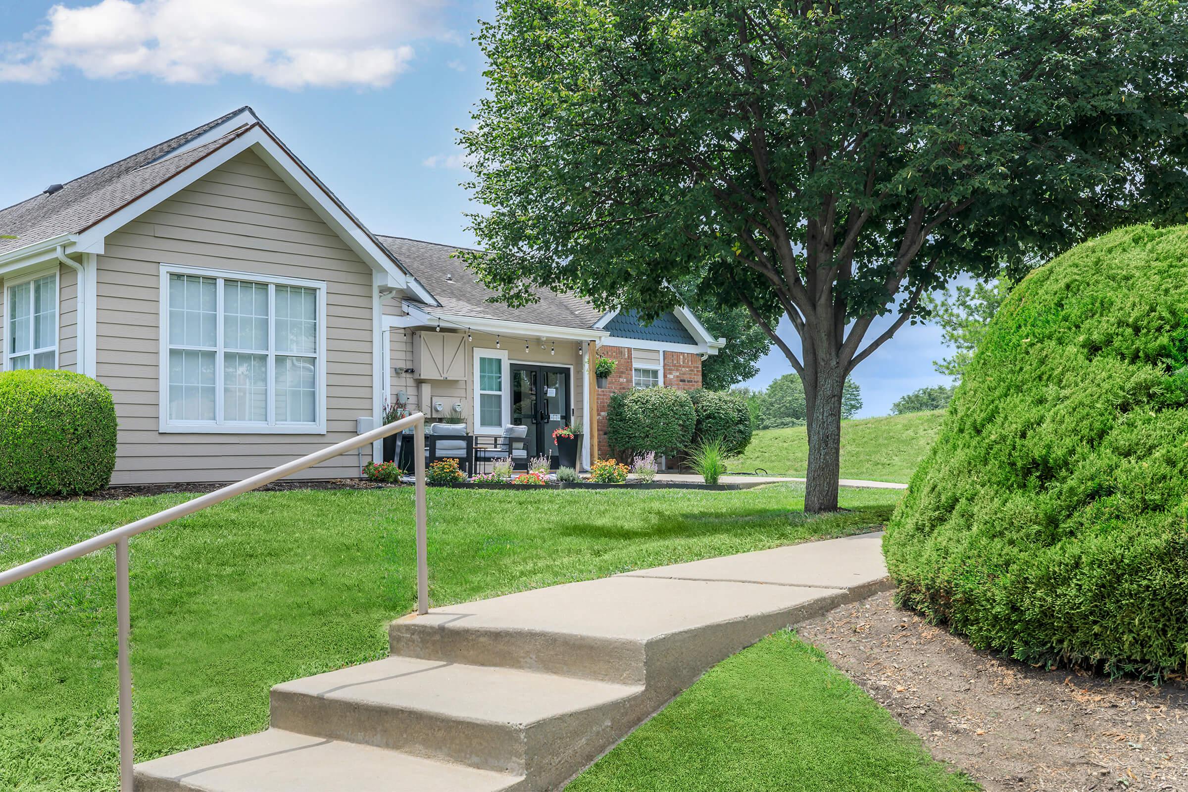 A well-maintained residential home with a light-colored exterior, large windows, and a welcoming front porch. The pathway leads up to the entrance, bordered by green lawns and neatly trimmed shrubs. A large tree provides shade in the yard, creating a pleasant and inviting atmosphere.