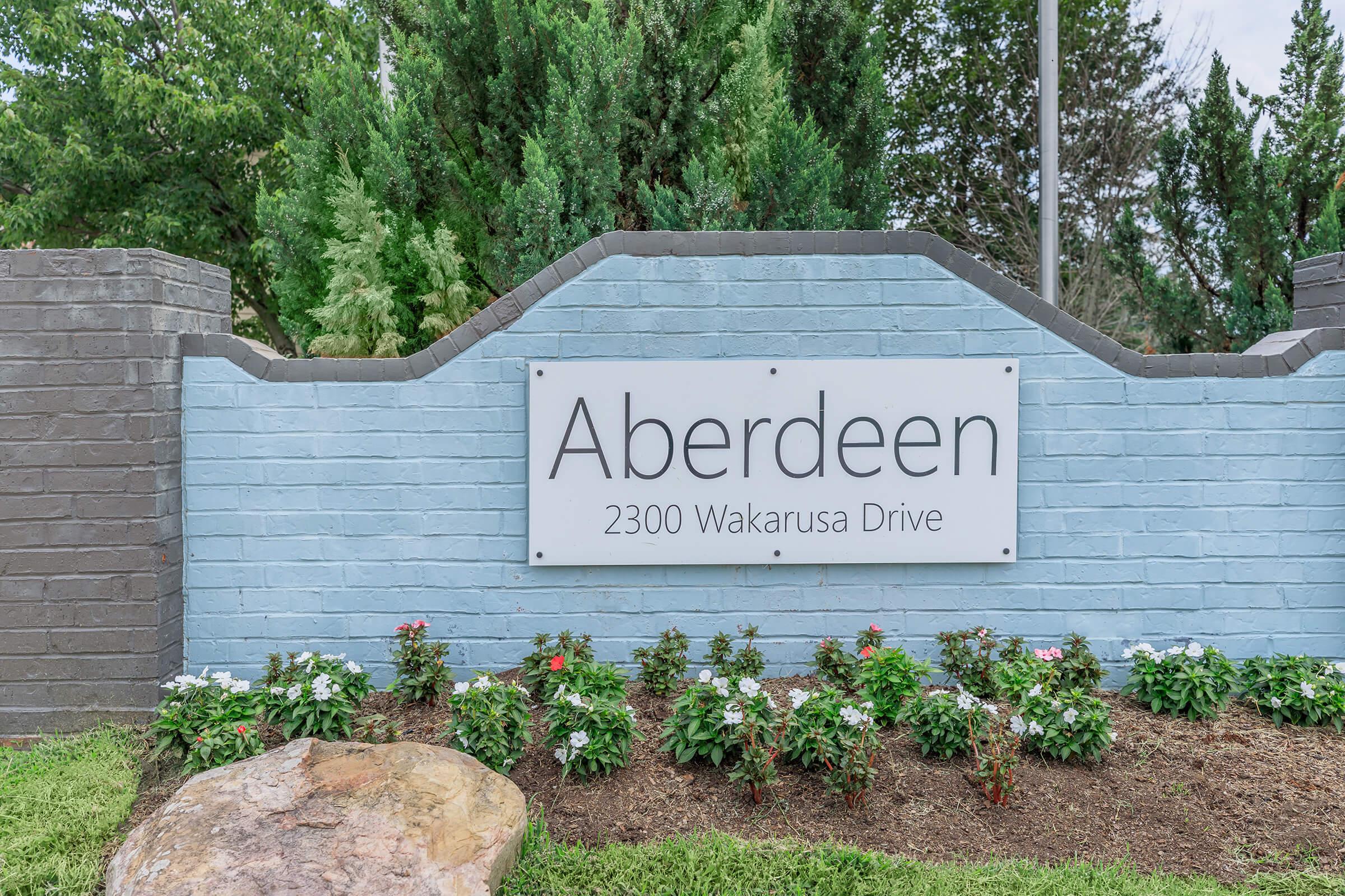 Sign for Aberdeen at 2300 Wakarusa Drive, featuring a light blue brick wall with green landscaping and flowers in front.