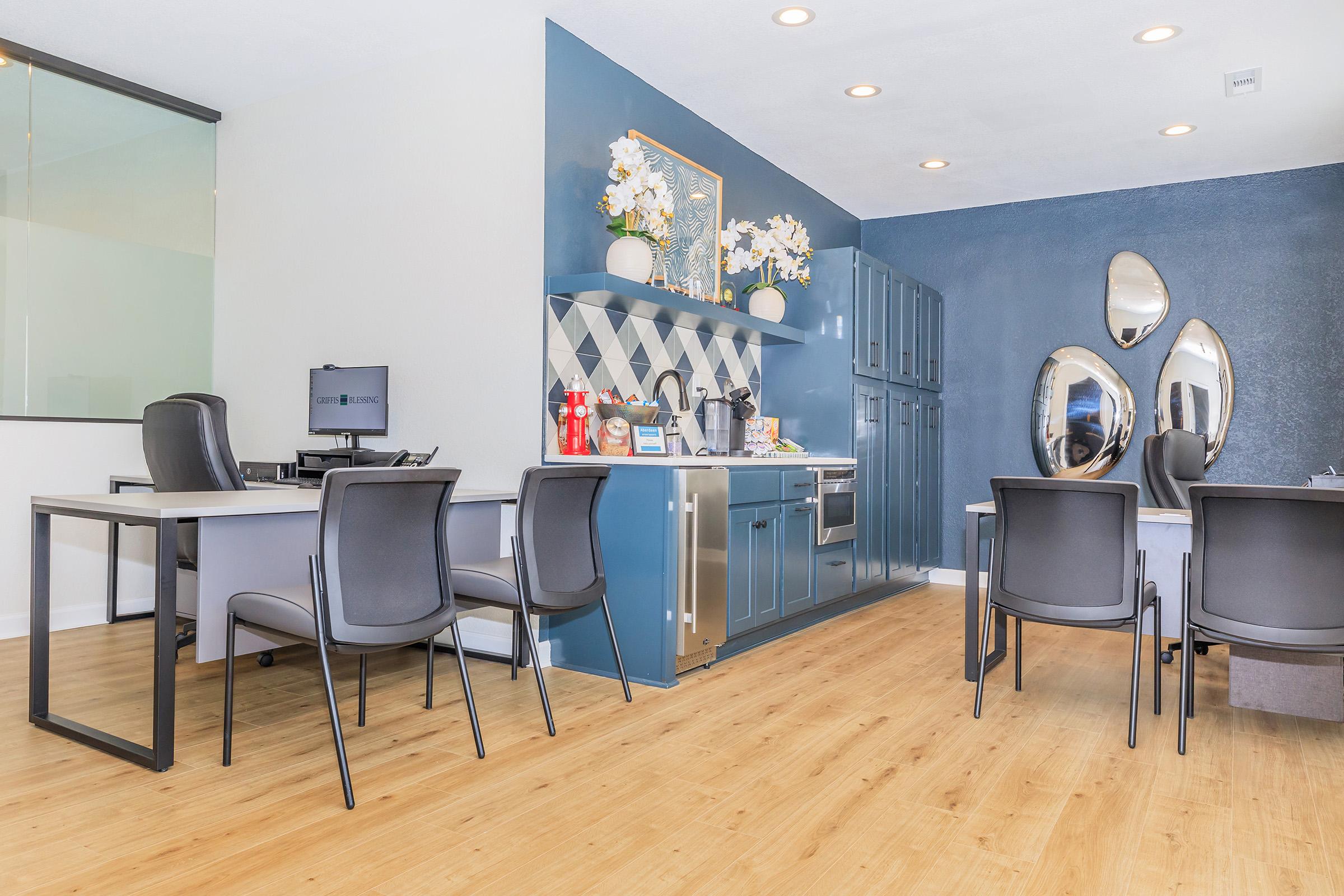 A modern office space featuring a kitchen area with blue cabinetry, decorative flowers, and mirrors on the wall. There are several black chairs around tables and a computer workspace visible, all set against a light wooden floor. The atmosphere is bright and inviting.