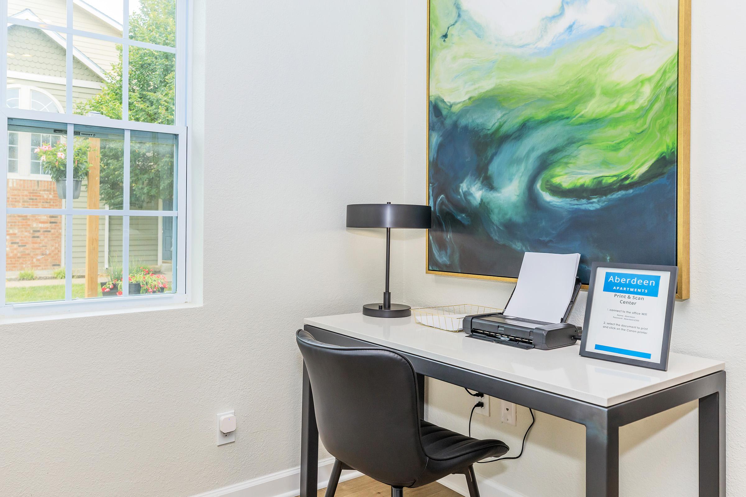 A bright and tidy workspace featuring a modern desk with a printer, a sleek black chair, and a decorative painting on the wall. A window allows natural light to fill the room, showcasing a view of greenery outside. A sign with "Aberdeen" is placed on the desk, adding a professional touch.