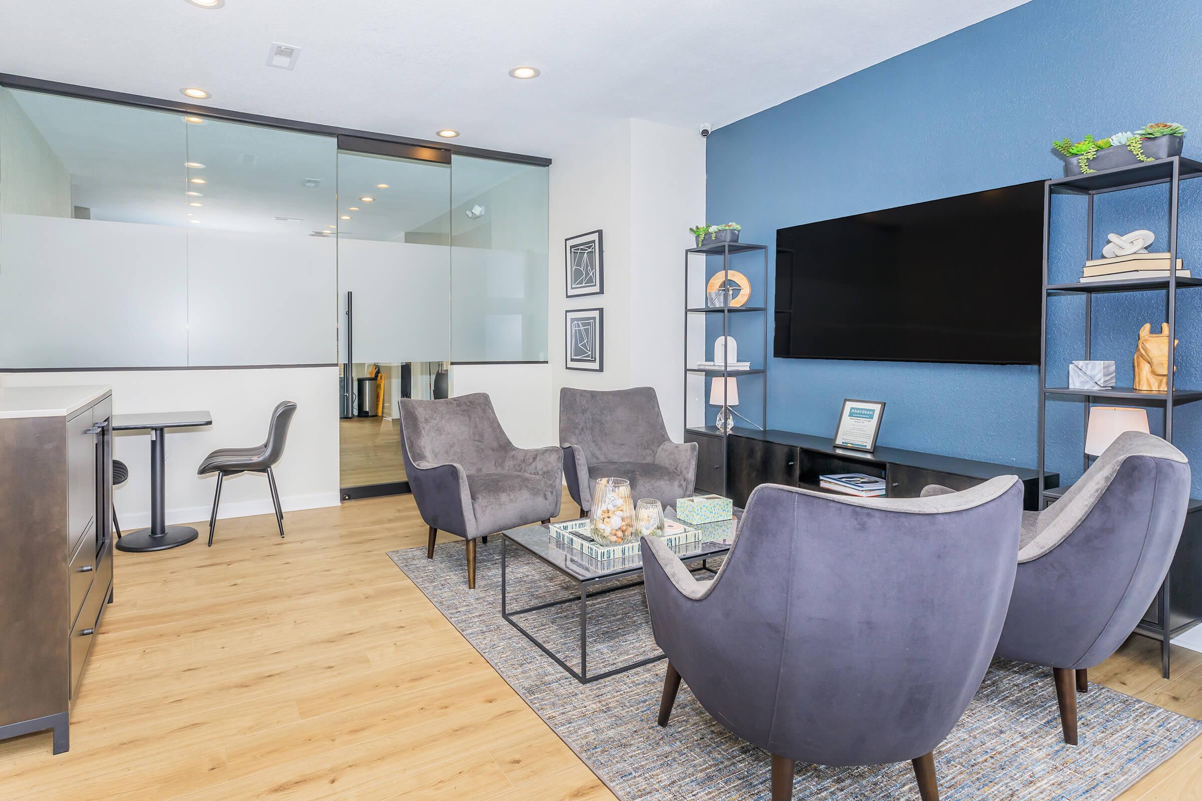 A modern living room featuring two stylish gray armchairs and a coffee table with decorative items. A flat-screen TV is mounted on the wall across from the seating area. The room has warm wood flooring, a glass wall that separates another space, and contemporary decor elements on shelves. Bright and inviting atmosphere.